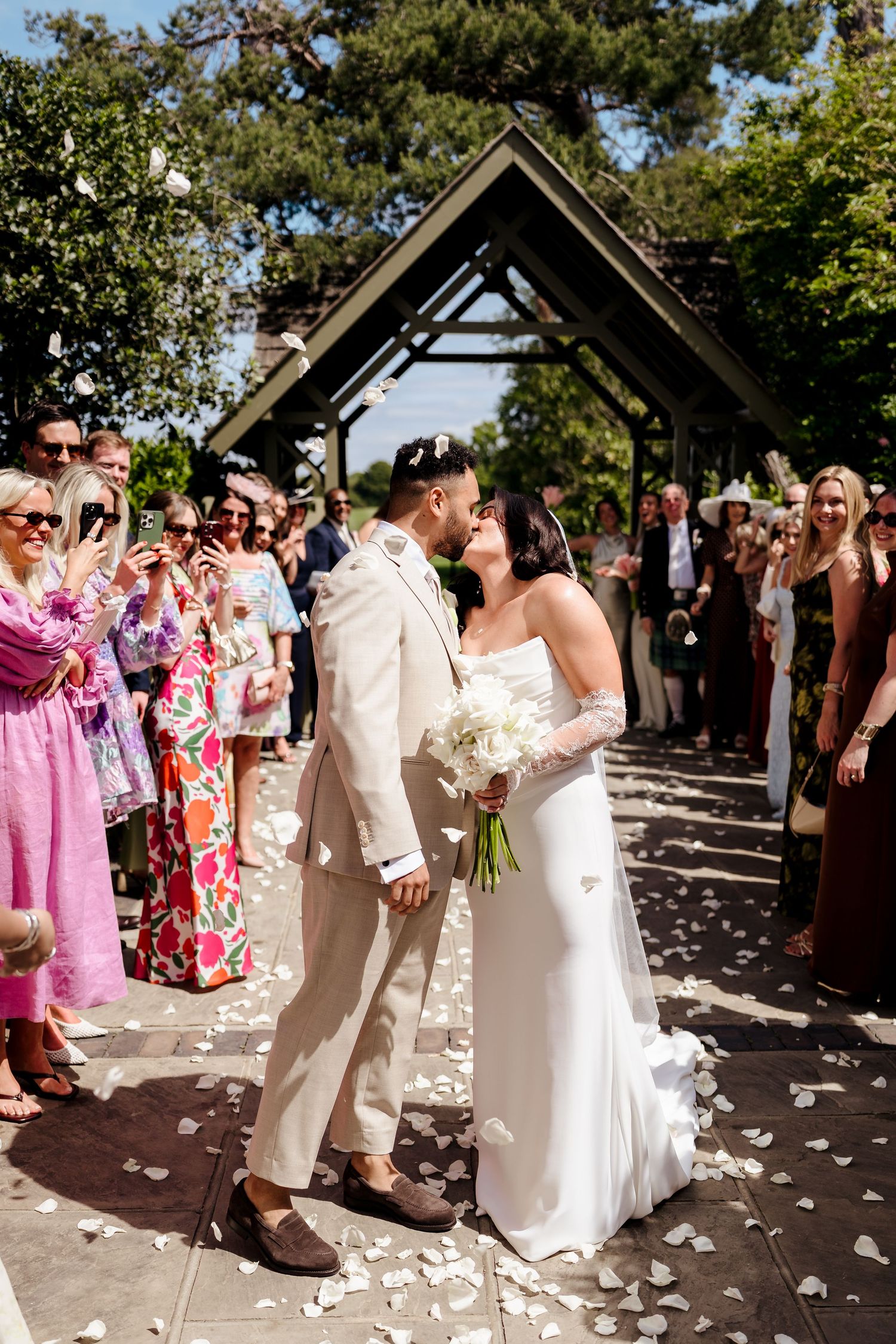 Bride and groom share a kiss while walking down the aisle as guests throw white flower petals at outdoor wedding.