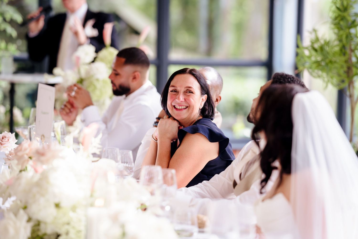 Smiling wedding guest in navy dress laughs at head table during elegant white floral wedding reception.