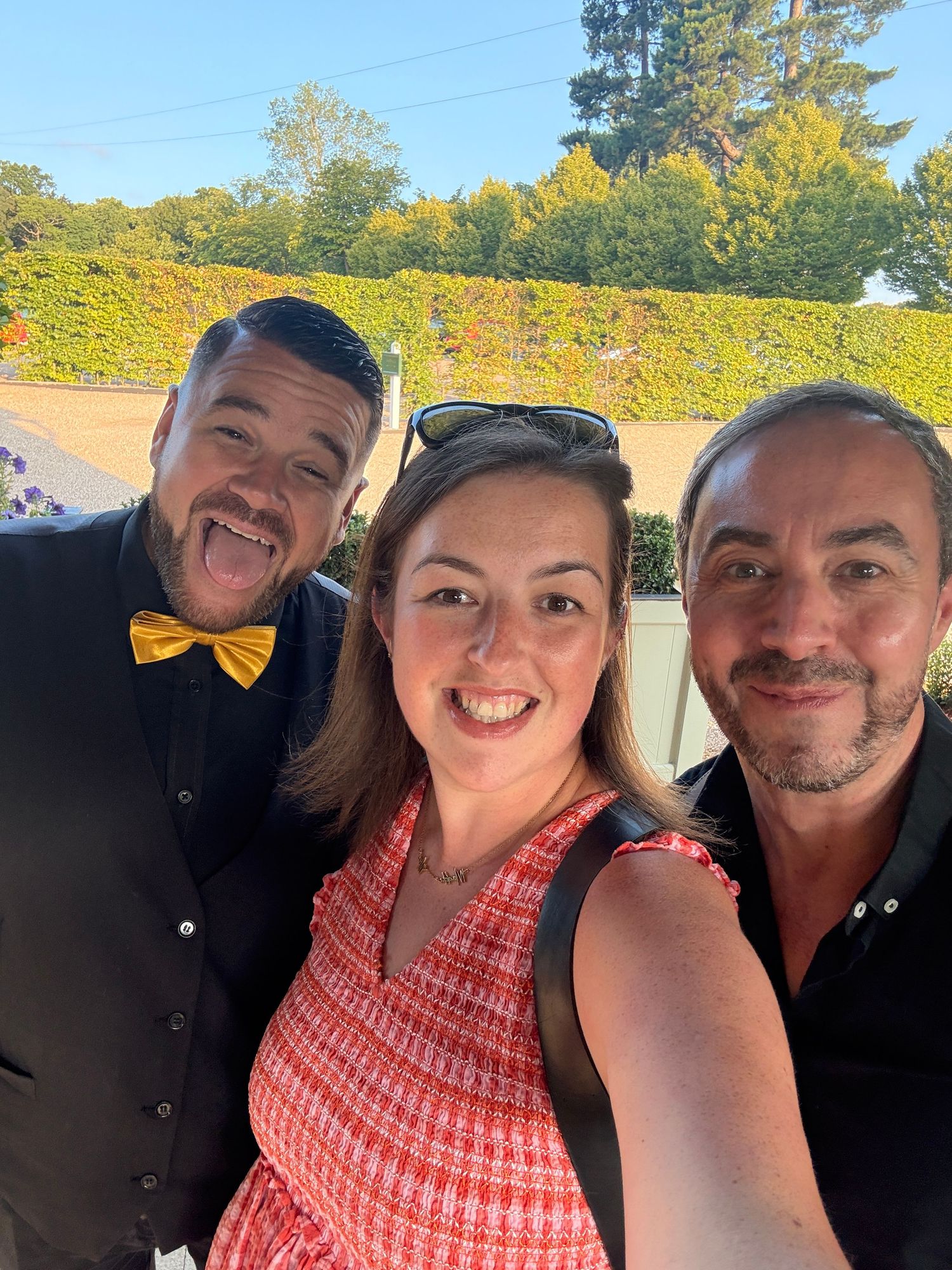 Three friends smiling for a selfie outdoors, man in yellow bow tie, woman in red dress, man in black shirt.
