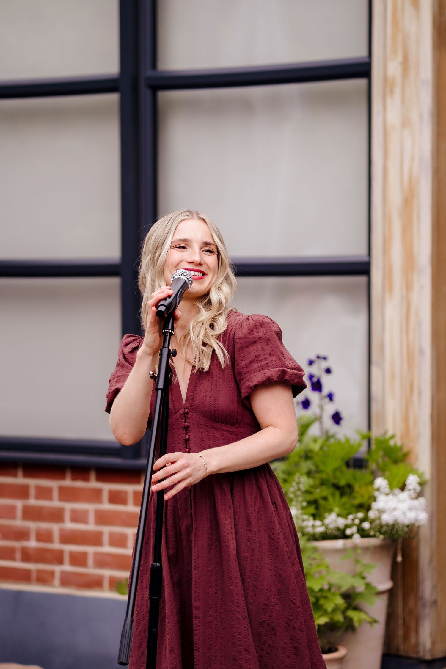 Blonde woman in burgundy dress singing at a microphone stand outdoors near brick wall and flowers.