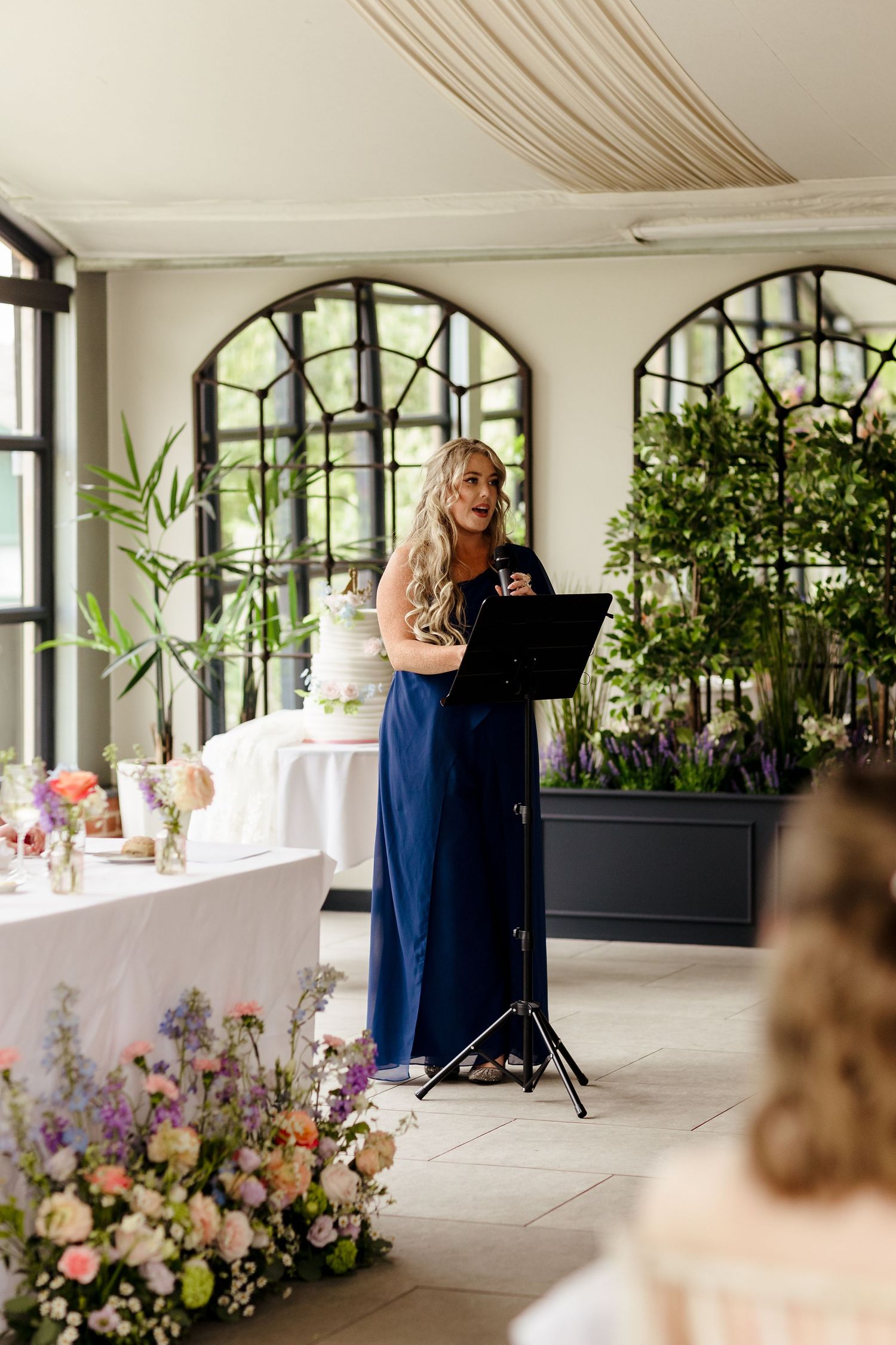 Bridesmaid in navy dress delivers a heartfelt speech at an elegant wedding reception.