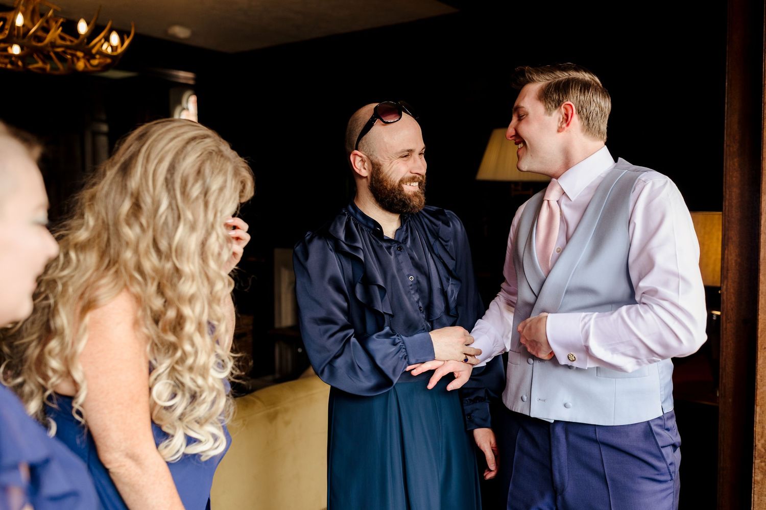 Groom in grey vest laughing with bearded officiant in navy robe during wedding preparations indoors.