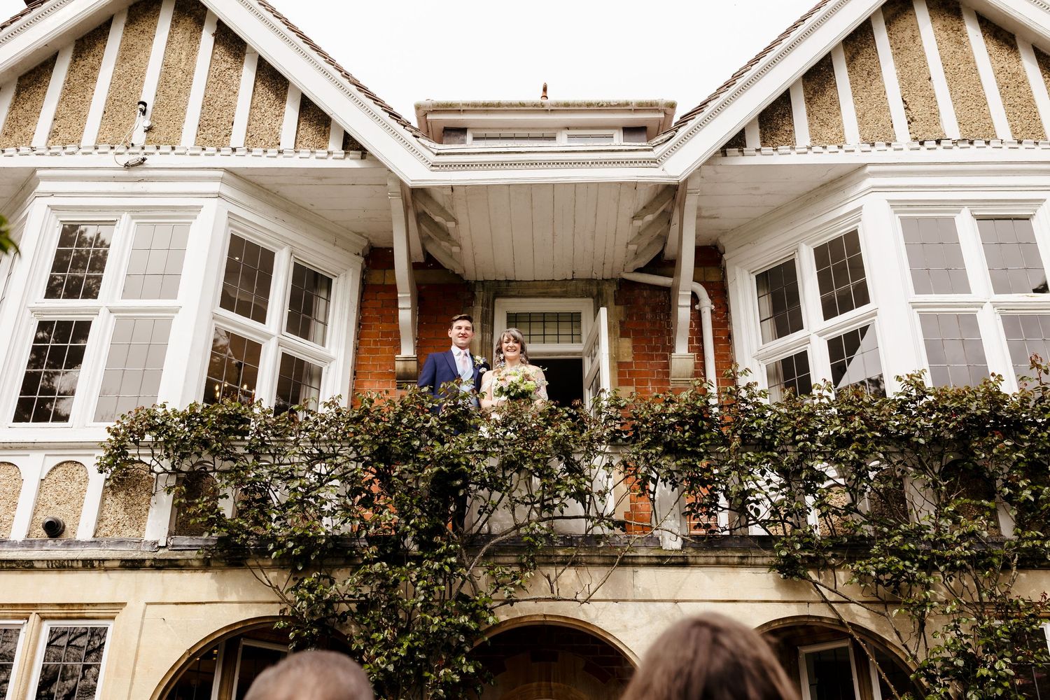 Bride and groom stand on ivy-covered balcony of elegant Tudor-style wedding venue.