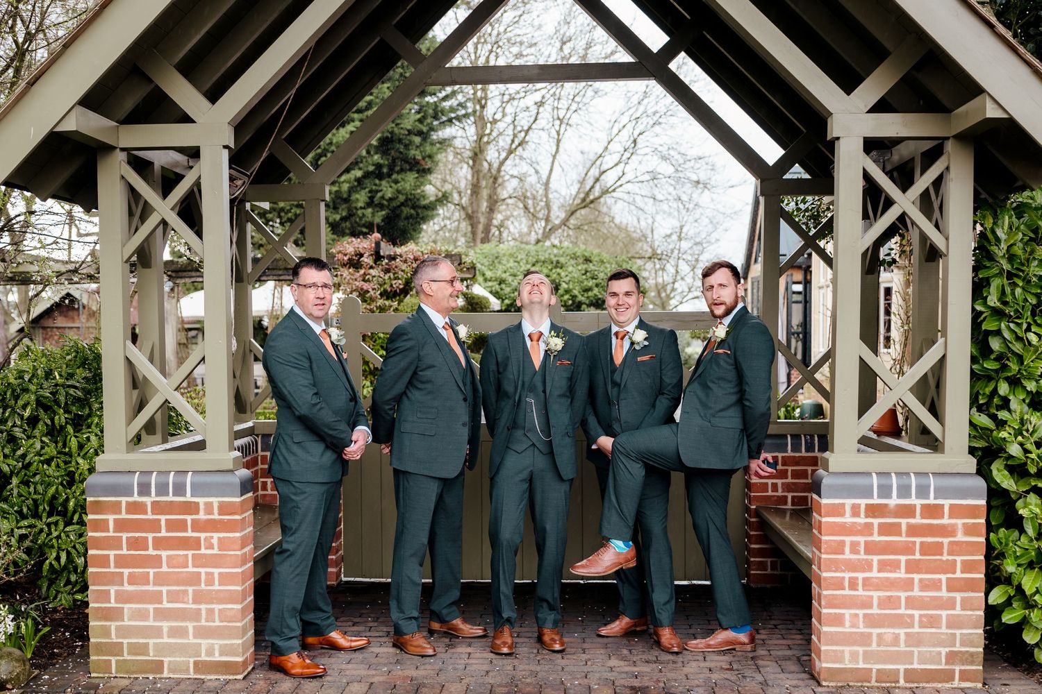 Groom and groomsmen in dark green suits with brown shoes posing under wooden gazebo at wedding venue.
