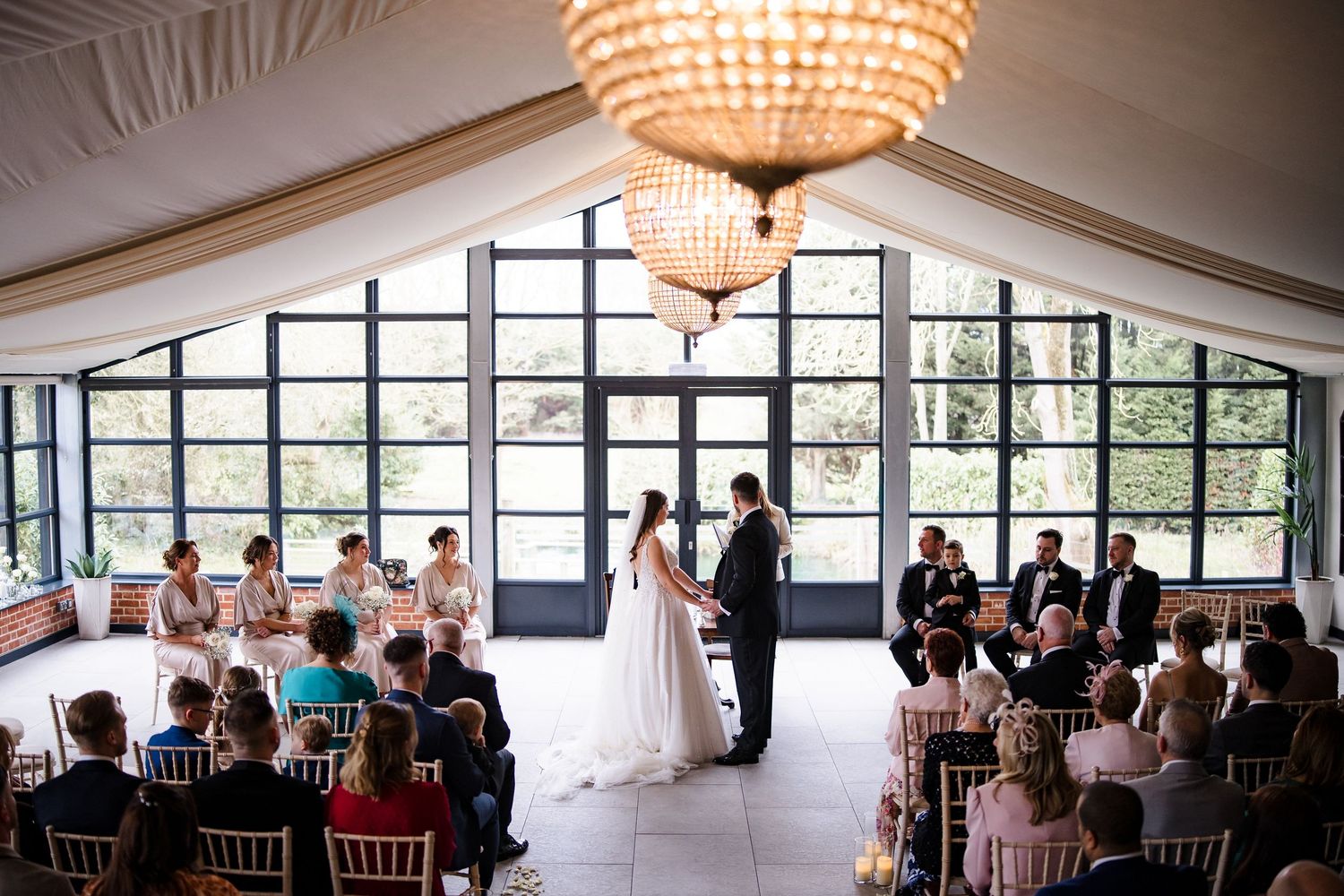 Elegant wedding ceremony in a bright venue with large windows, couple at altar with guests seated.