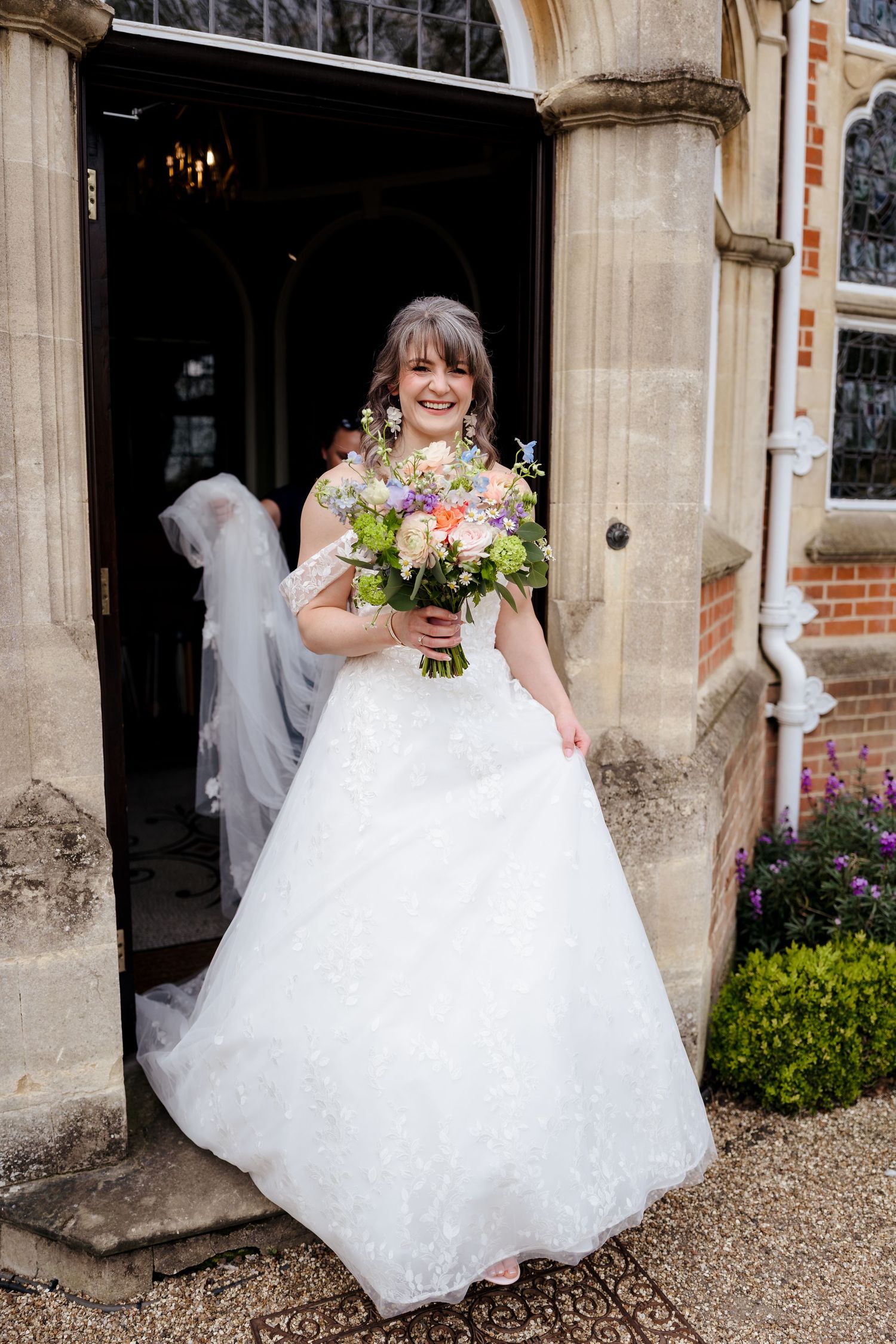 Smiling bride in lace wedding dress holding colorful bouquet standing at church doorway.