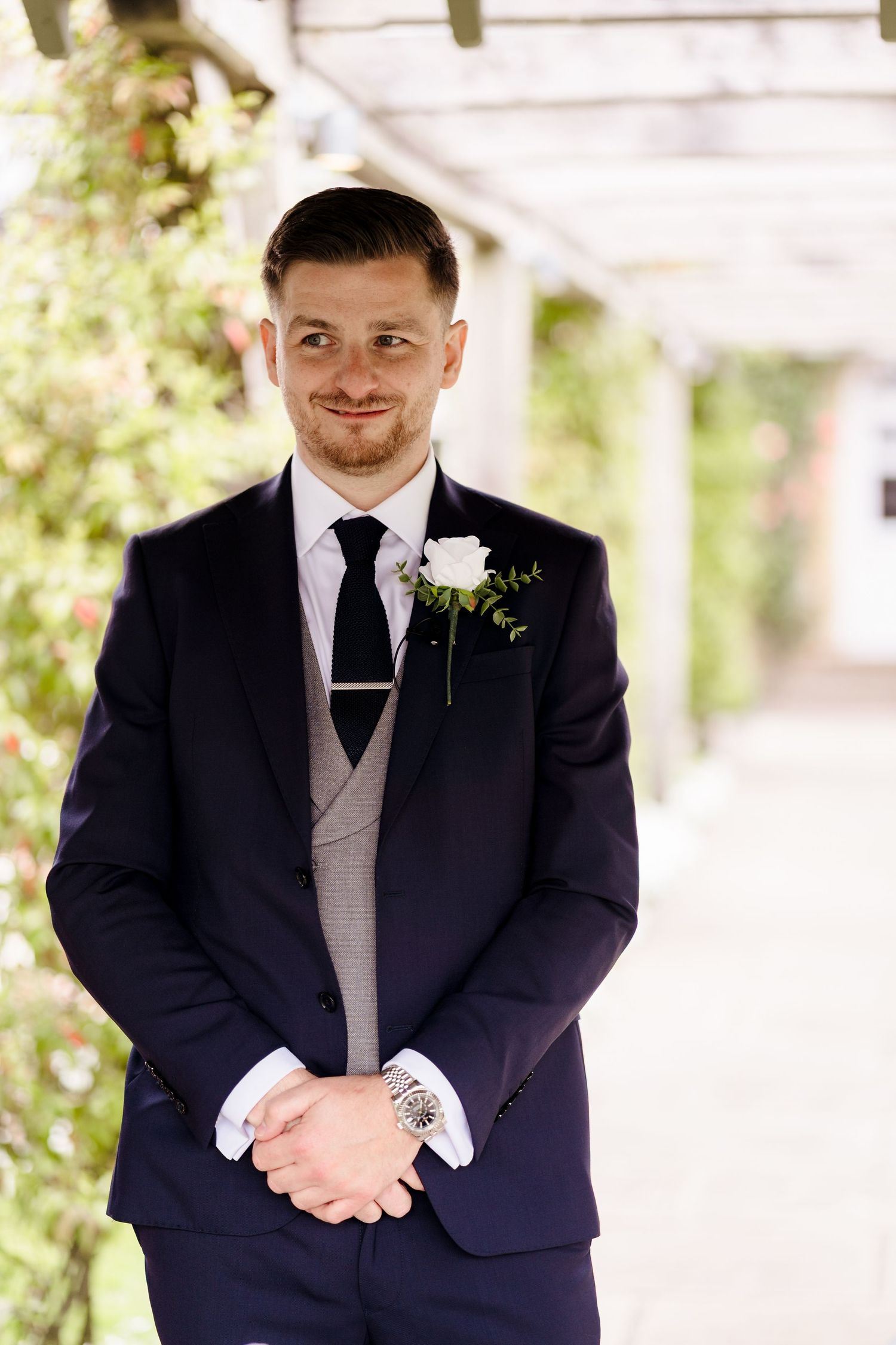 Smiling groom in navy suit with white rose boutonniere and black tie, standing outdoors at a wedding venue.