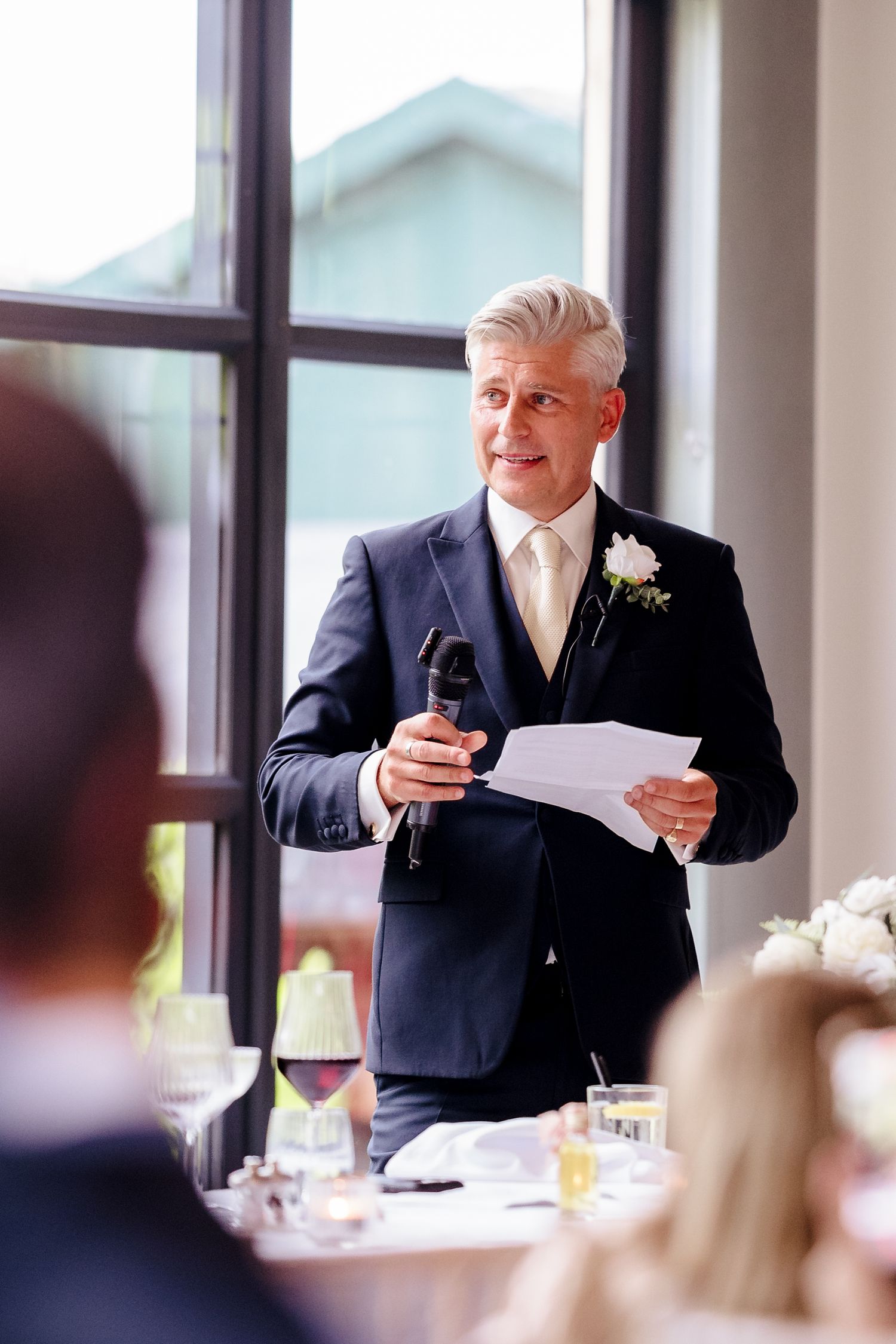 Elegant silver-haired man in navy suit delivers heartfelt wedding speech holding microphone and notes at reception.