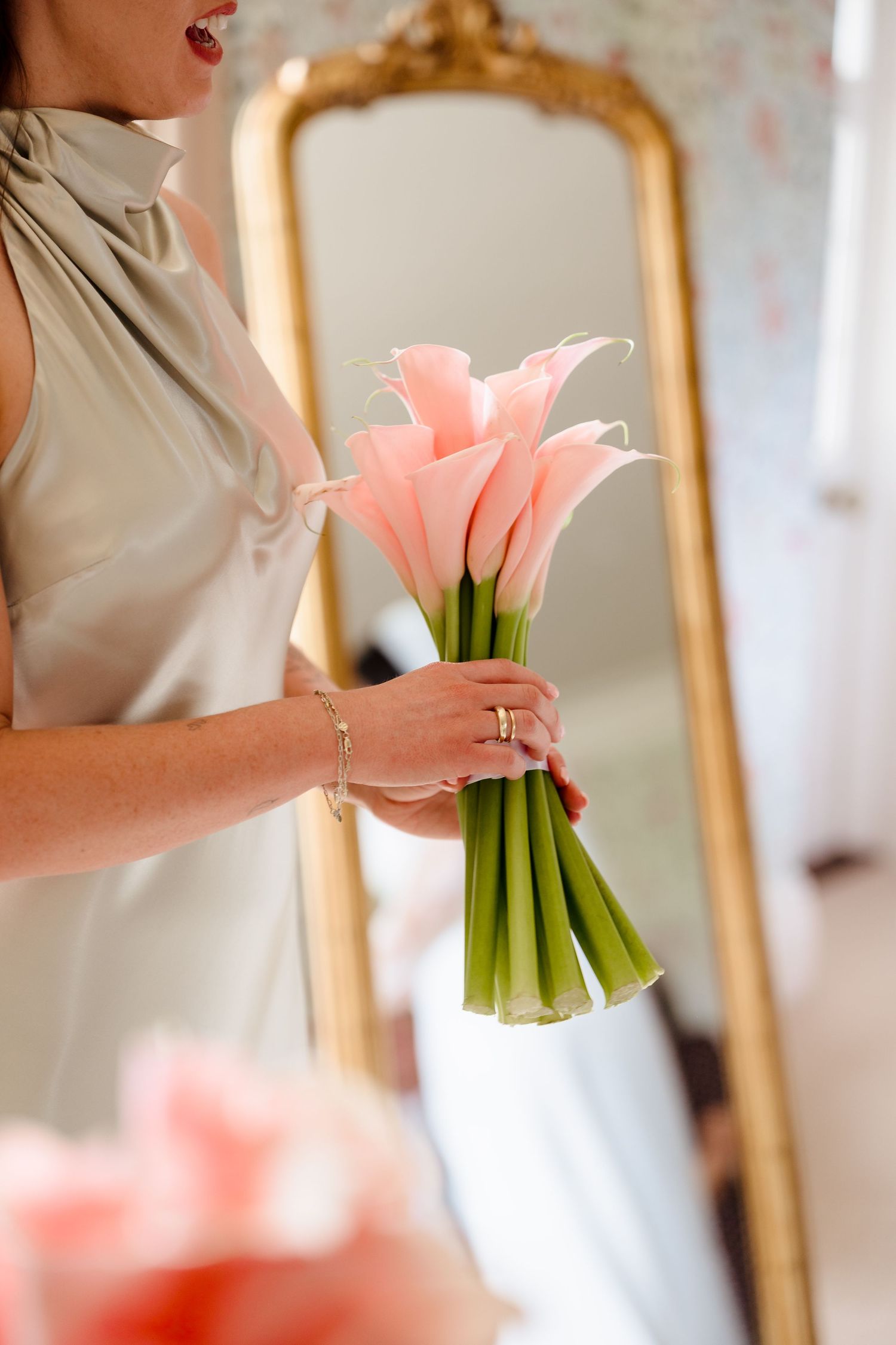 Bride holding a bouquet of pink calla lilies in front of a gold ornate mirror.