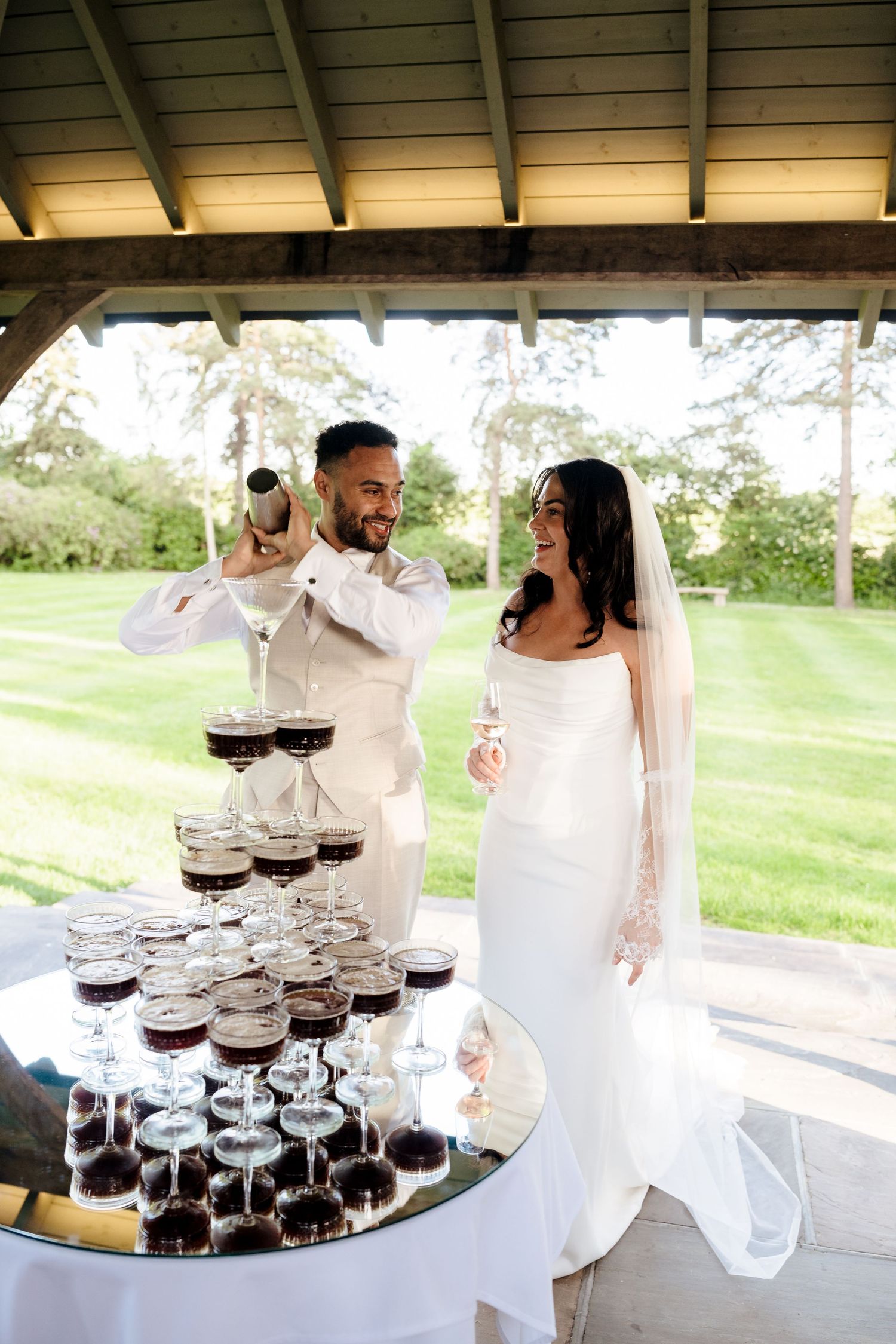 Groom pouring champagne tower while bride laughs beside him at outdoor wedding reception.