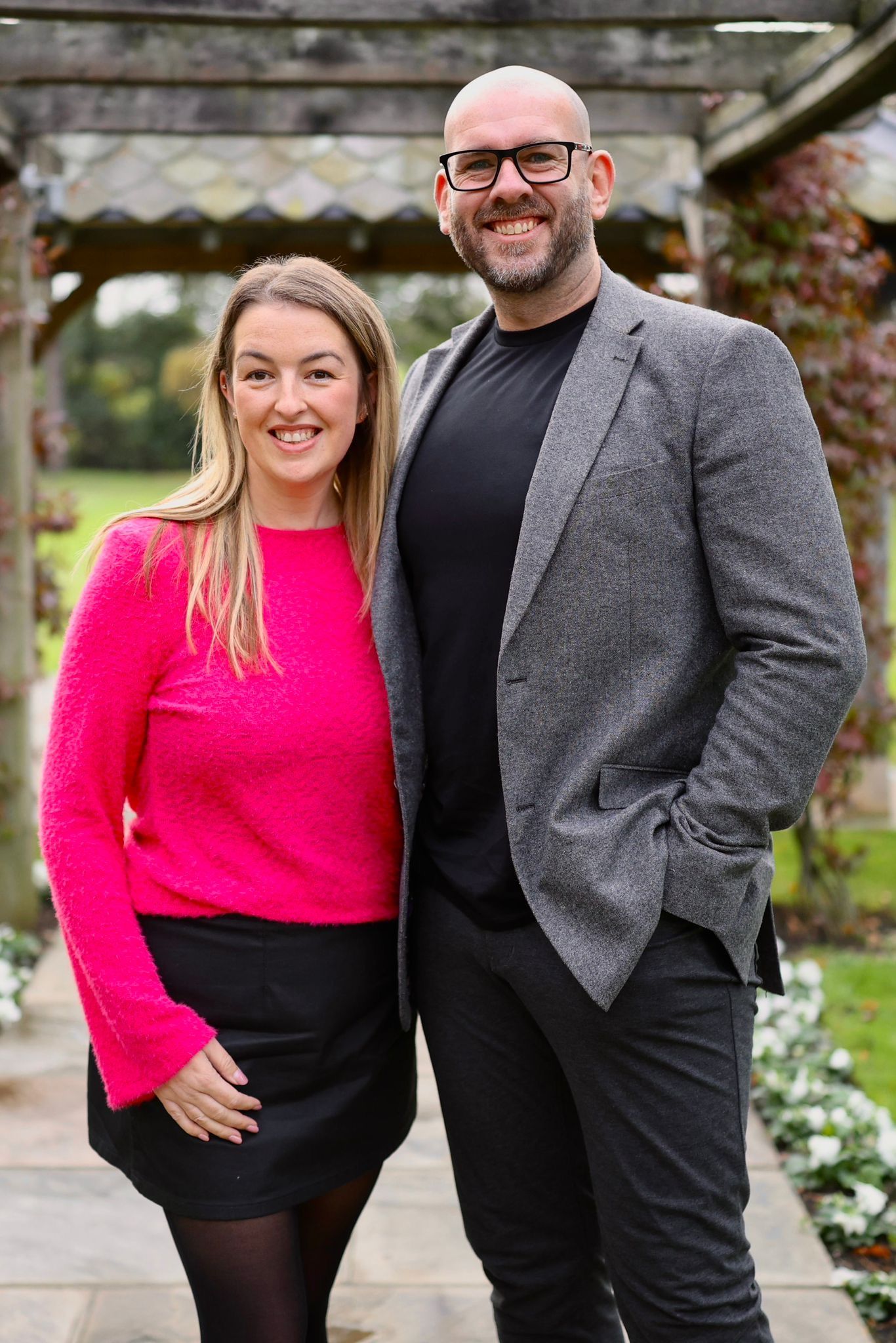 A smiling couple posing outdoors, woman in pink sweater and man in grey blazer, standing near a wooden pergola.