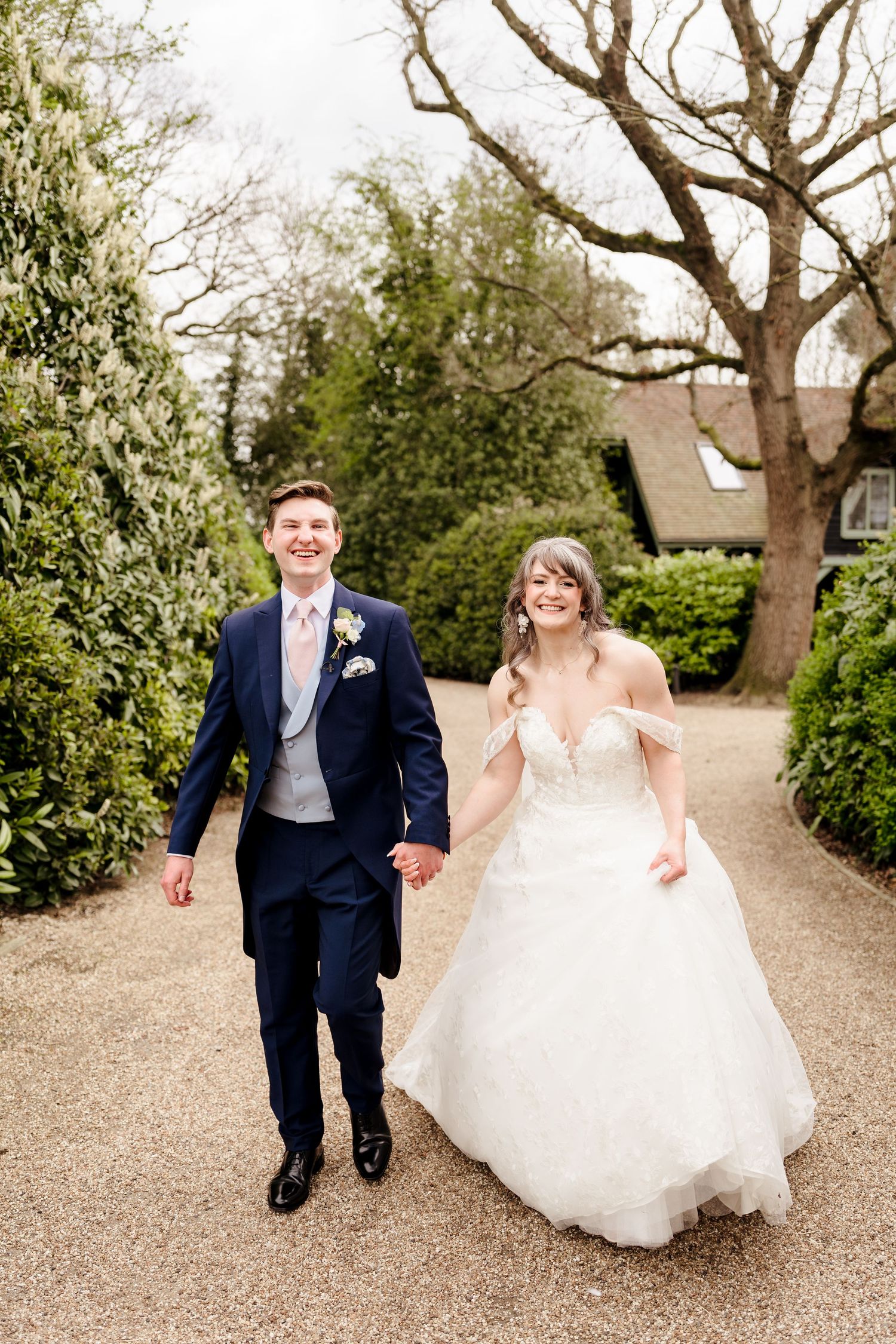A smiling bride and groom walk hand in hand along a gravel path surrounded by lush greenery on their wedding day.
