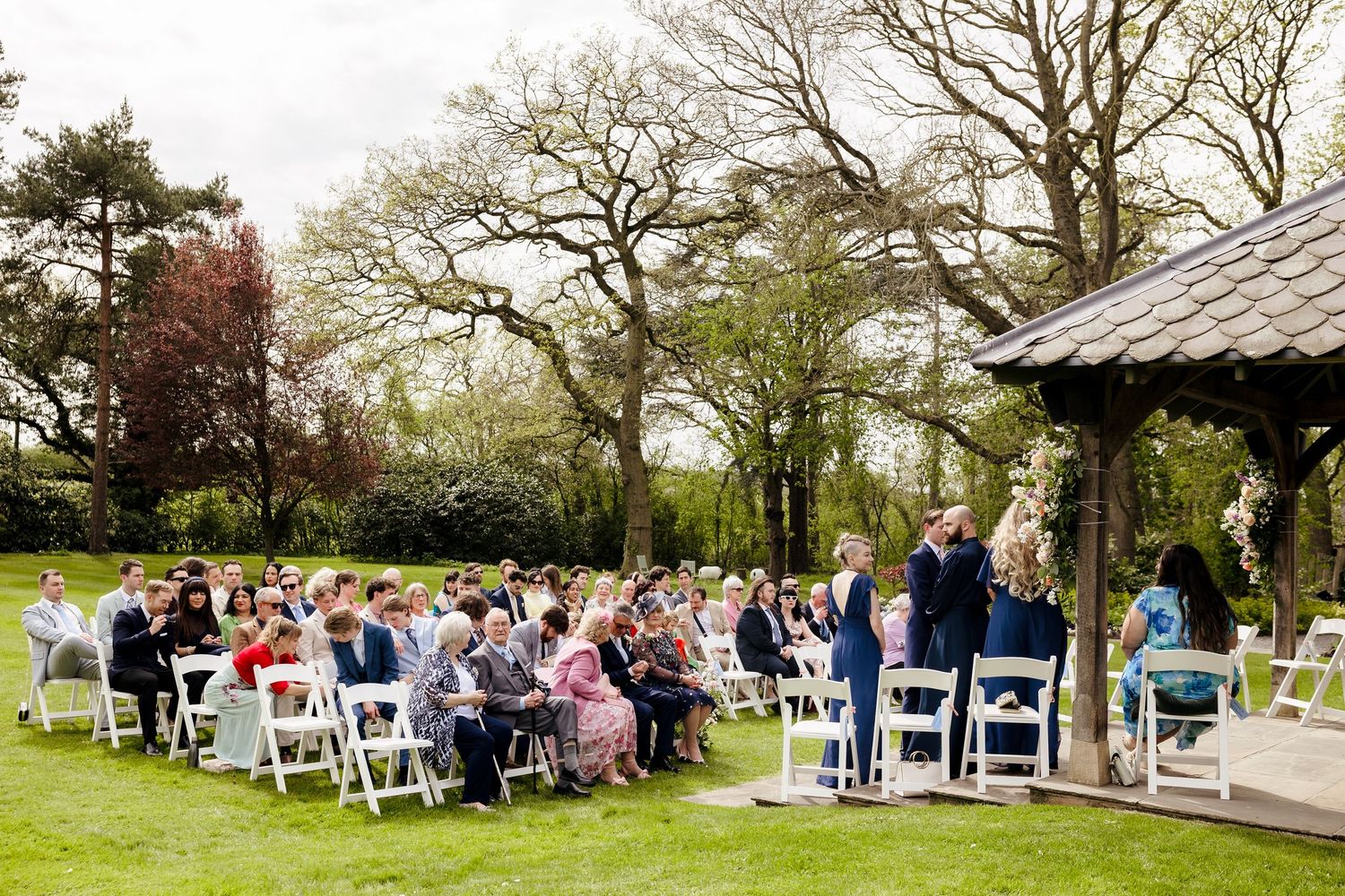 Outdoor wedding ceremony with guests seated on white chairs in a lush garden with tall trees and a gazebo.