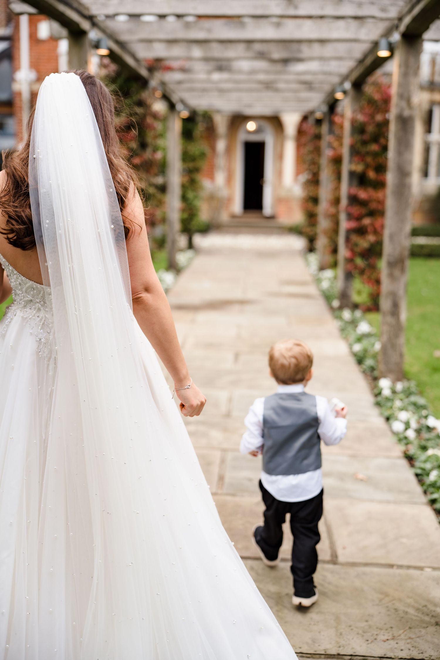 Bride in white gown and veil walks with young boy in vest along garden pathway toward elegant venue entrance.