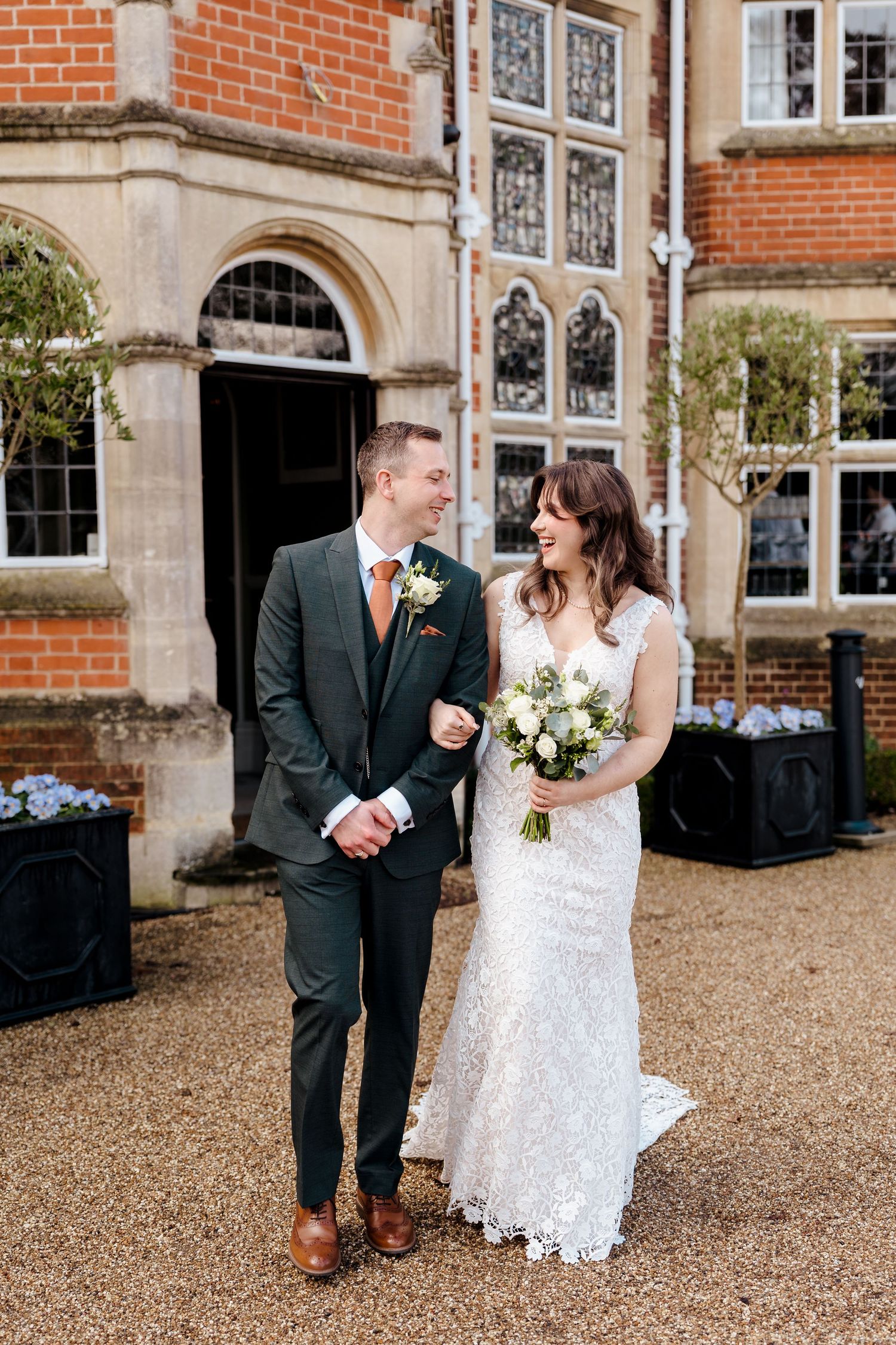 Smiling bride and groom pose outside a grand red brick manor house on their wedding day.