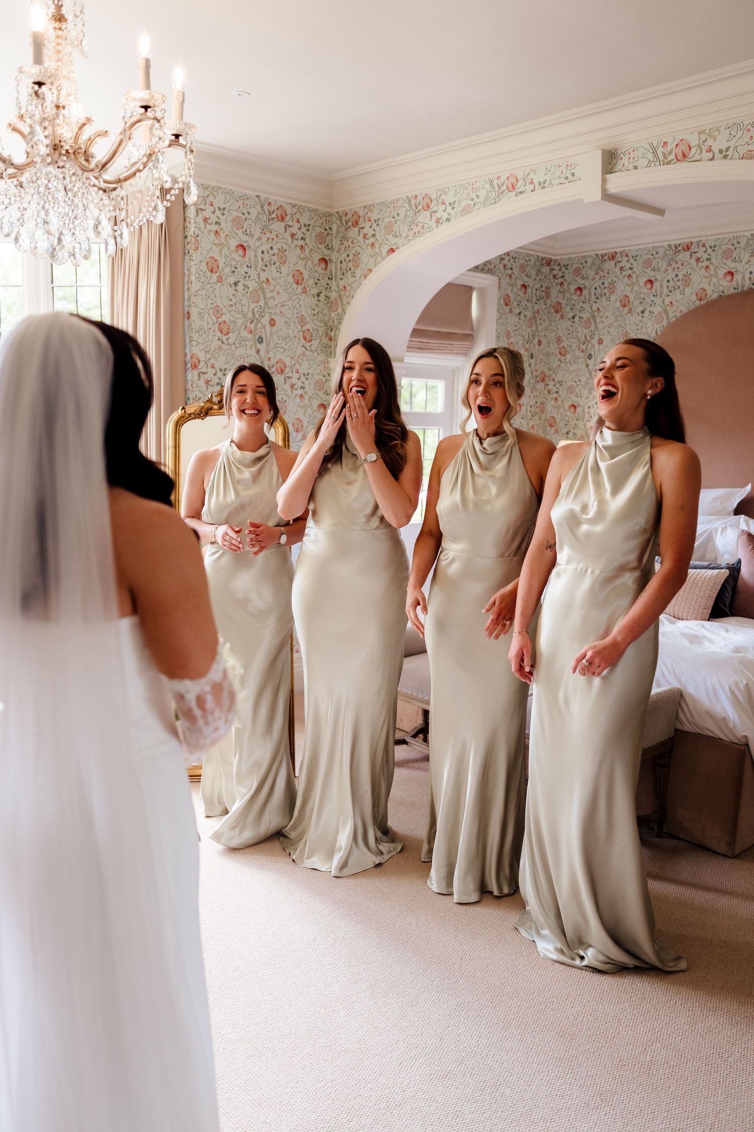 Bridesmaids in sage satin gowns react with joy seeing bride in wedding dress in floral wallpapered room.