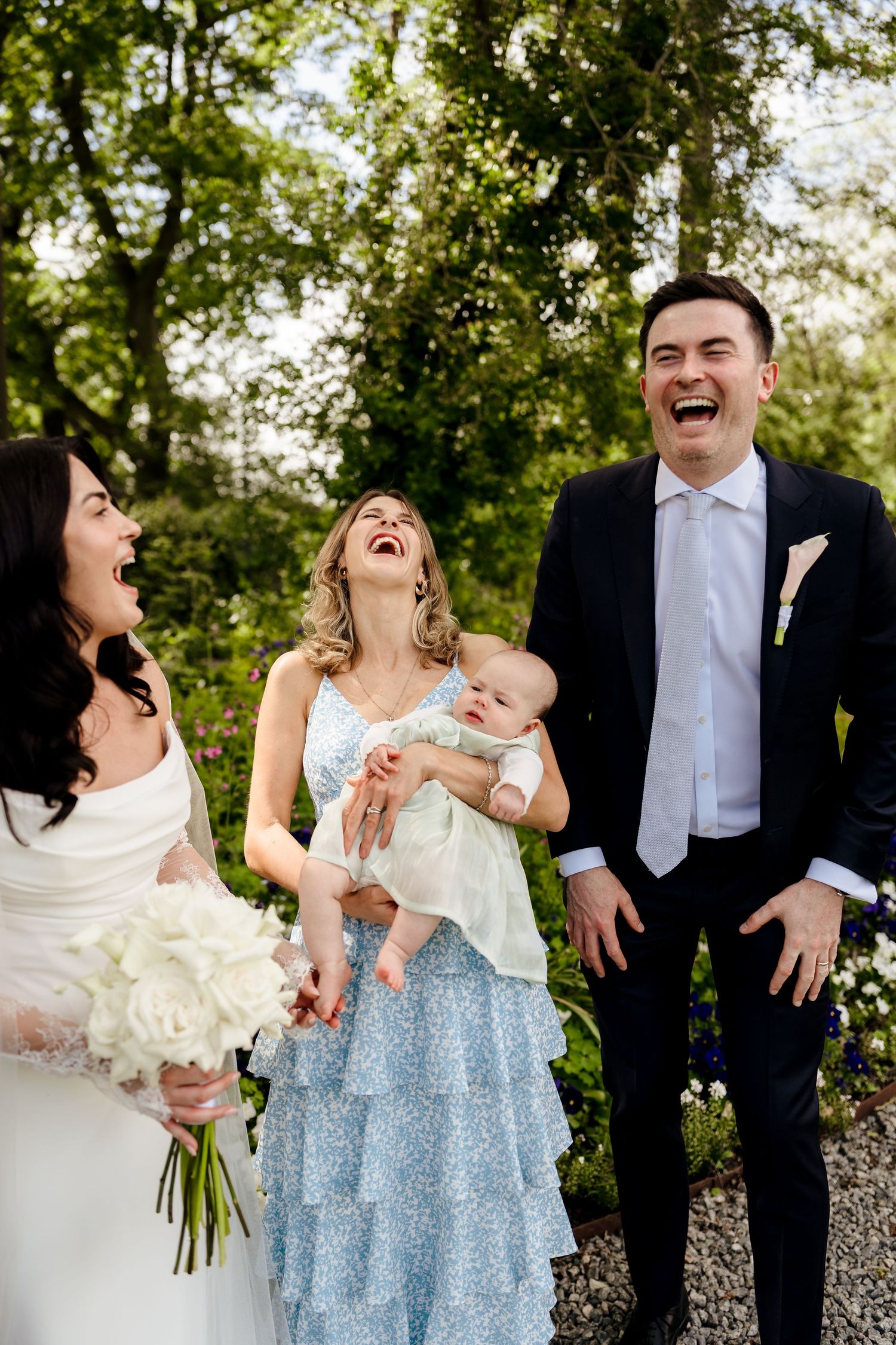 Bride, groom, and guests laughing joyfully outdoors at a wedding celebration with a baby.