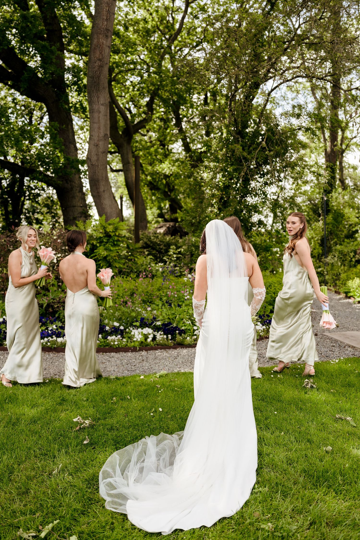 Bride in white gown with long veil stands with bridesmaids in champagne satin dresses in lush garden.