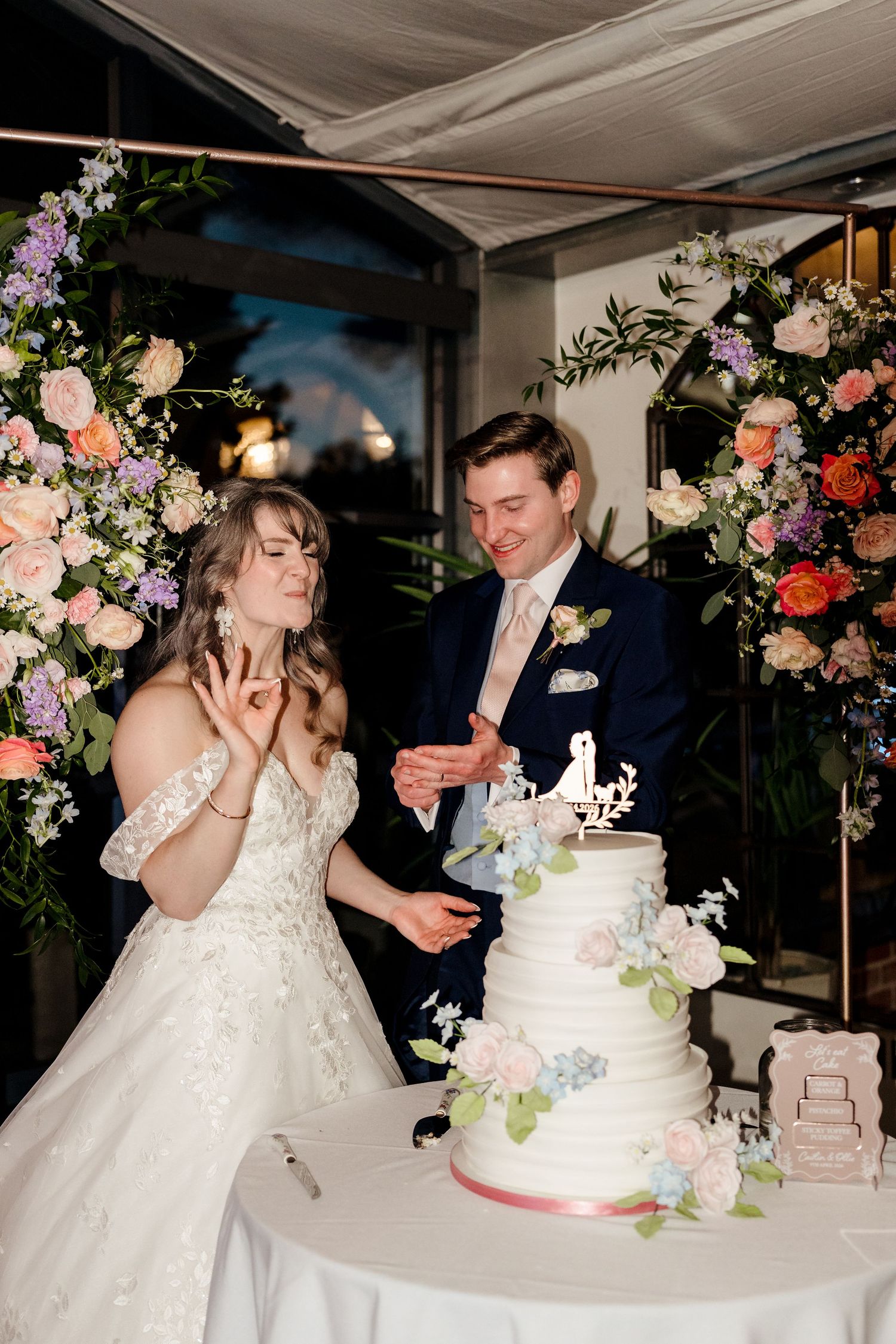 Bride and groom cut tiered white wedding cake adorned with flowers under floral arch.