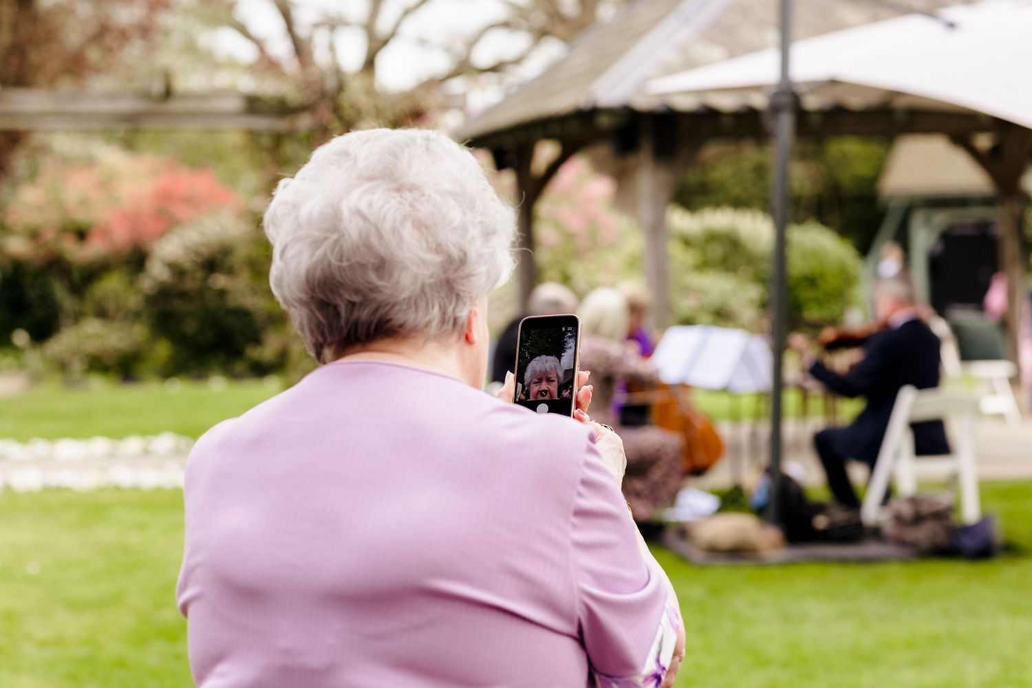 Elderly woman in pink photographing musicians at an outdoor garden gazebo event with her smartphone.