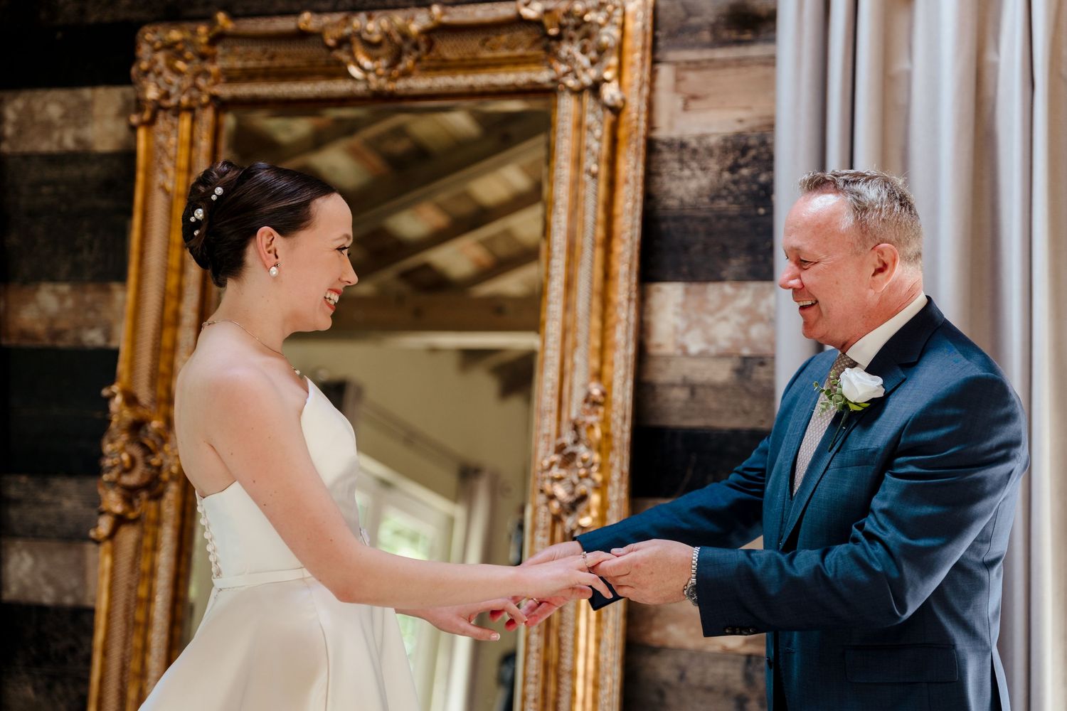 Bride and groom exchanging rings indoors, smiling joyfully, with an ornate gold mirror and rustic wood wall backdrop.