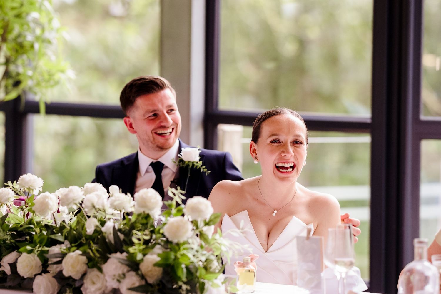 Happy bride and groom laugh together at wedding reception table adorned with white floral arrangements.