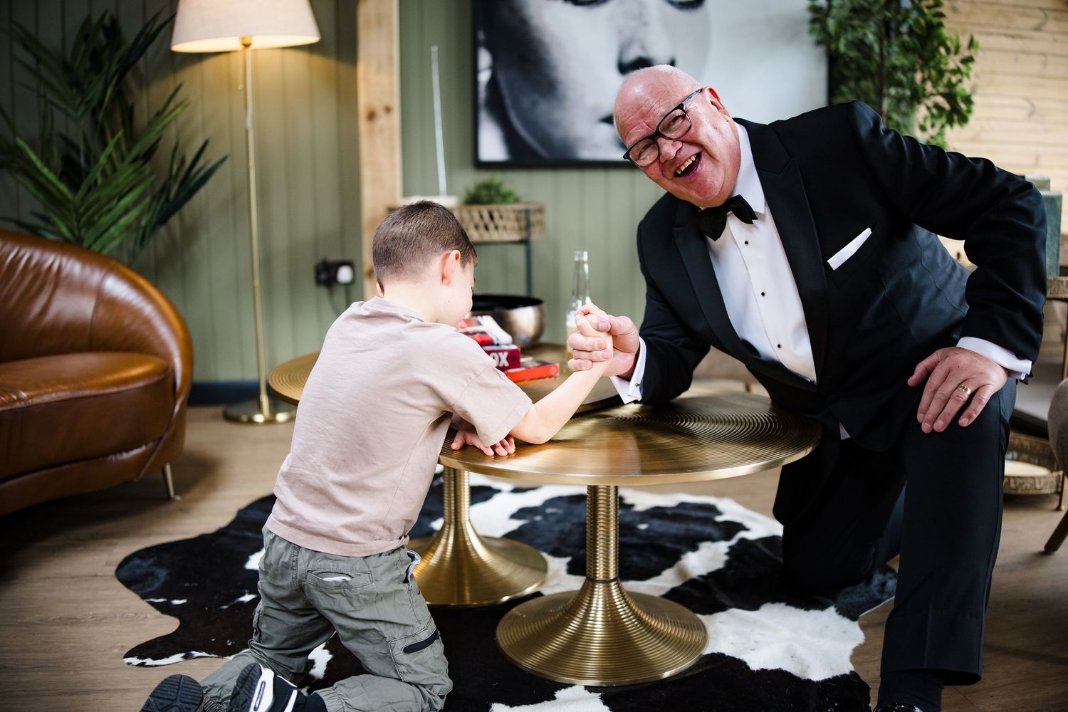 An elderly man in a tuxedo arm wrestling with a young boy at a gold table, both smiling and laughing.
