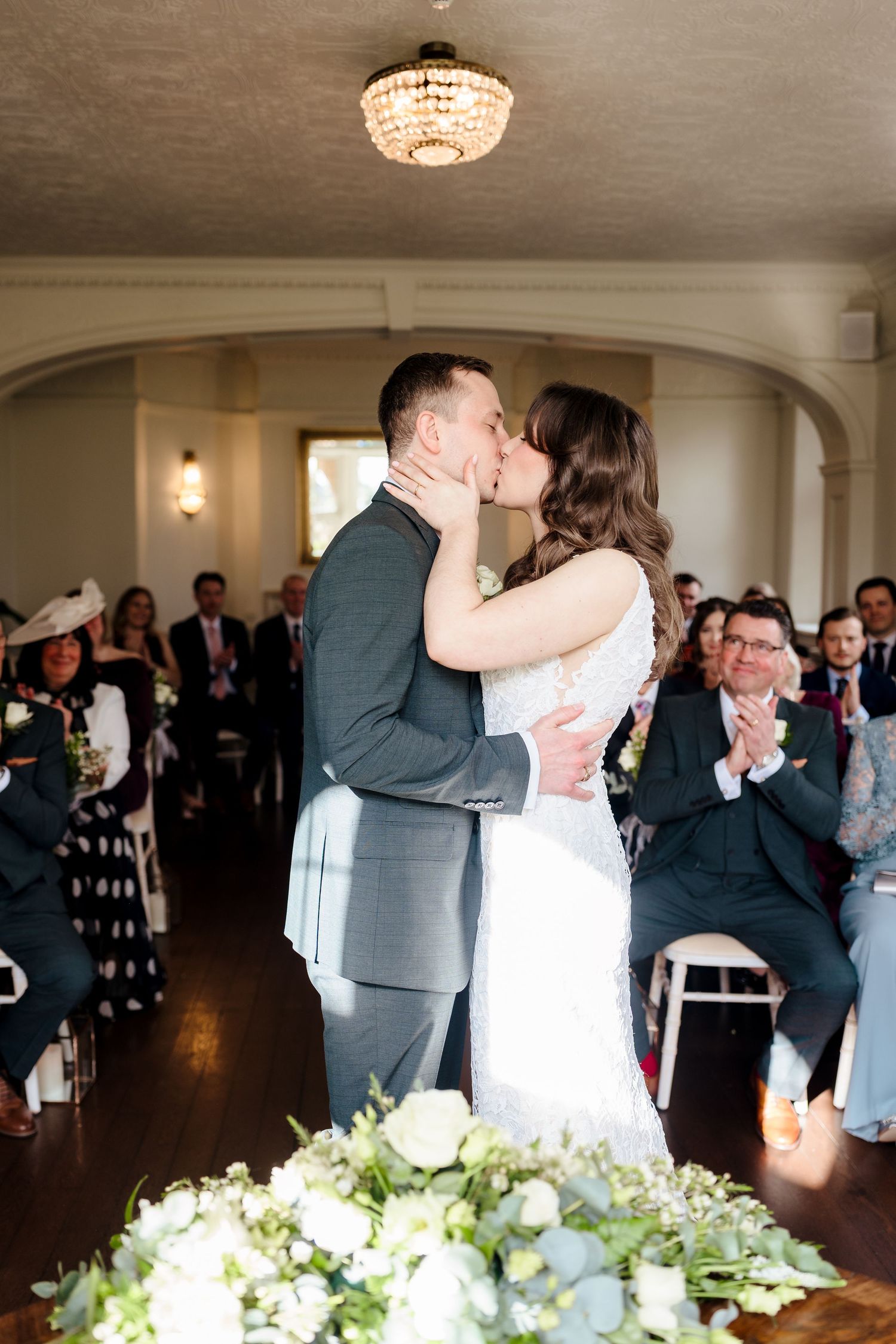 Bride and groom share first kiss at wedding ceremony while guests applaud in elegant white decorated venue.