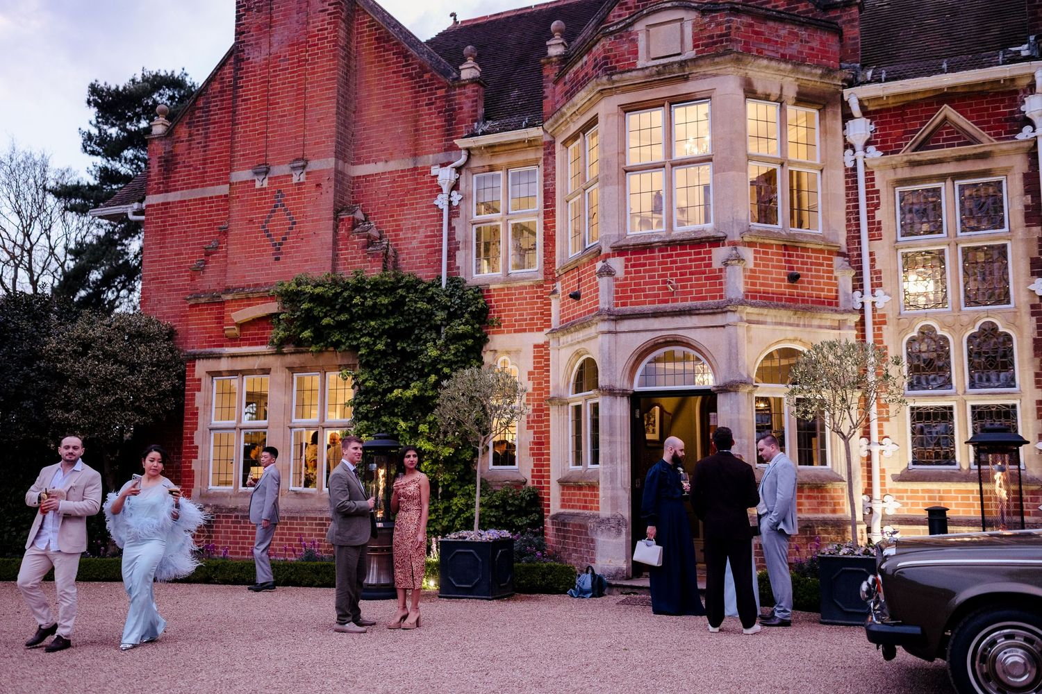 Wedding guests gathering outside a grand red brick manor house at dusk with vintage car.