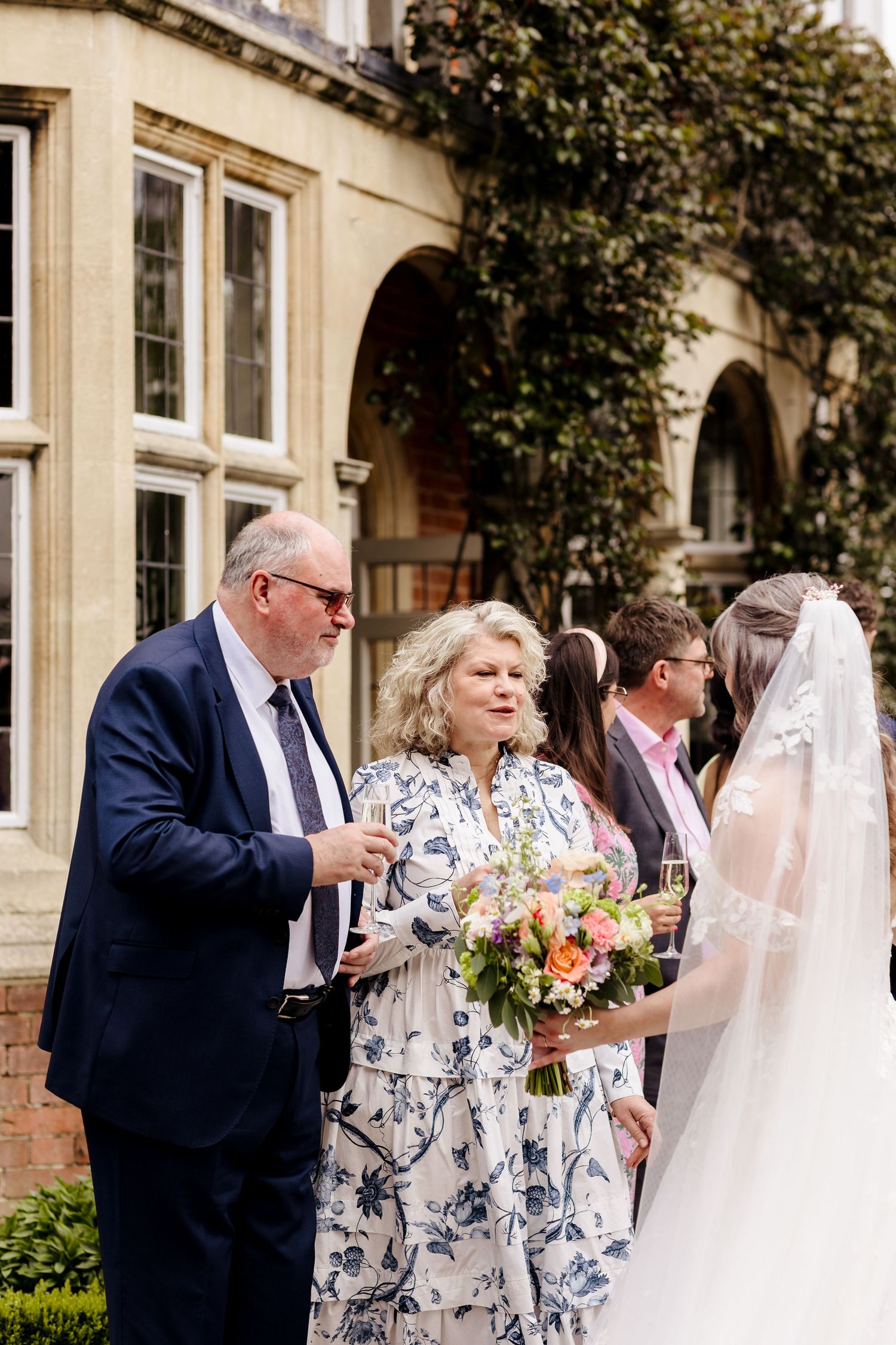 Bride in lace veil holds colorful bouquet while chatting with guests outside stone wedding venue.