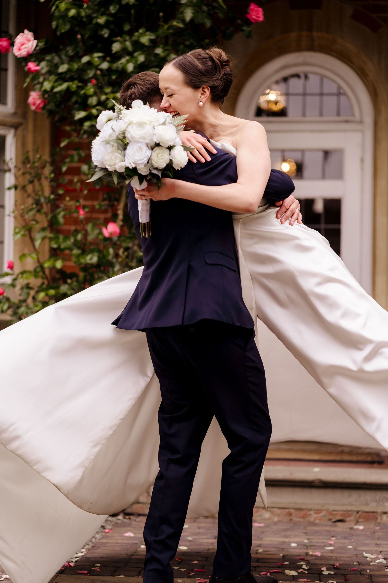 Bride in strapless white gown embraces partner in navy suit holding white bouquet outside wedding venue.