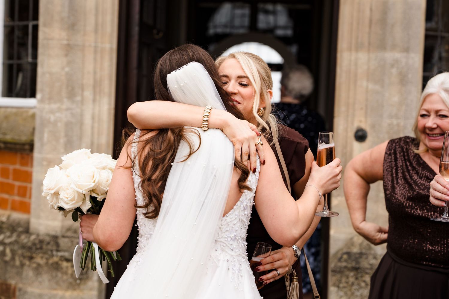 Bride in white gown and veil hugging a guest at a wedding reception outdoors.