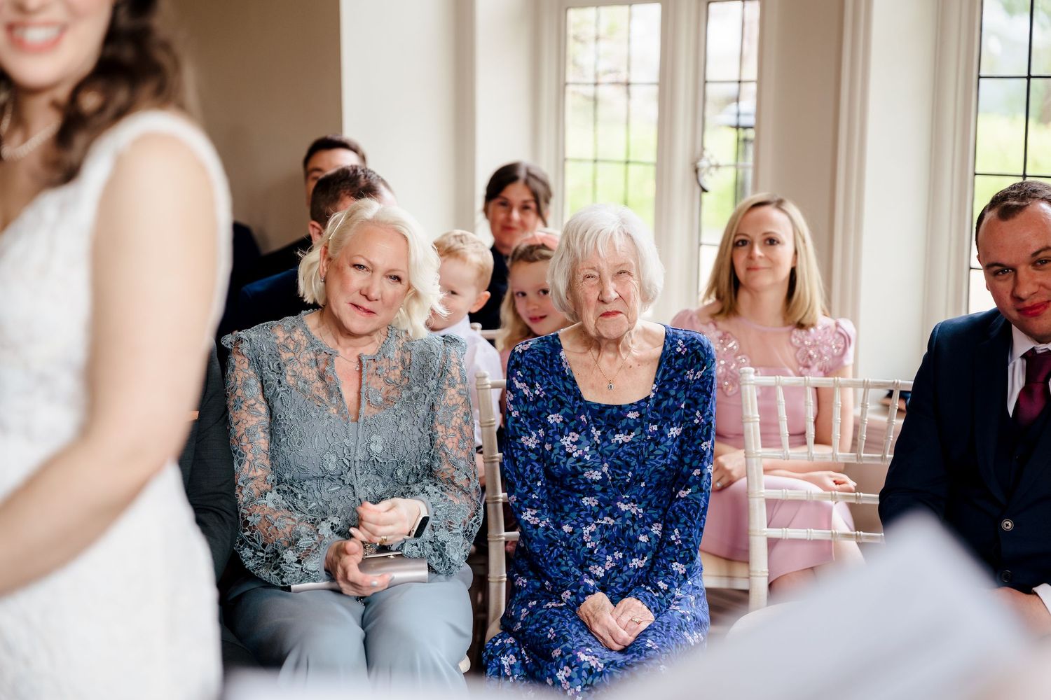 Guests seated at a wedding ceremony, two women in blue lace dresses watching attentively in a bright elegant venue.