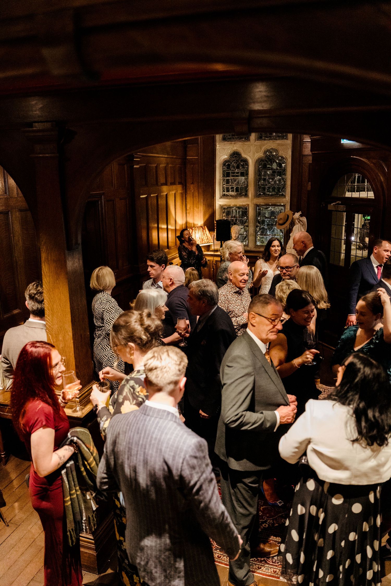 Wedding guests mingle in an elegant wood-paneled venue with stained glass windows during a reception.