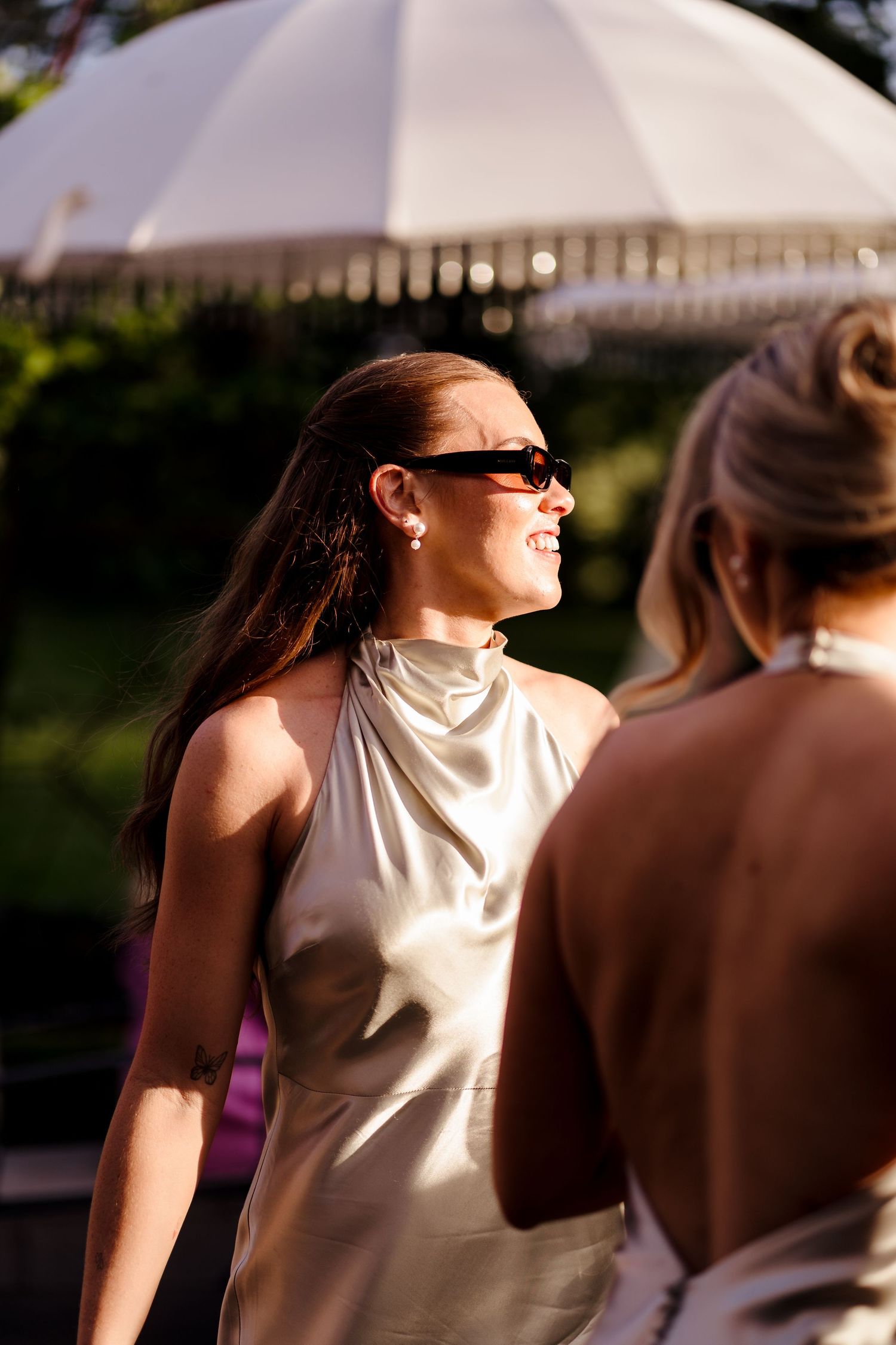 Elegant bridesmaid in silver halter dress and sunglasses smiling at golden hour outdoor wedding.