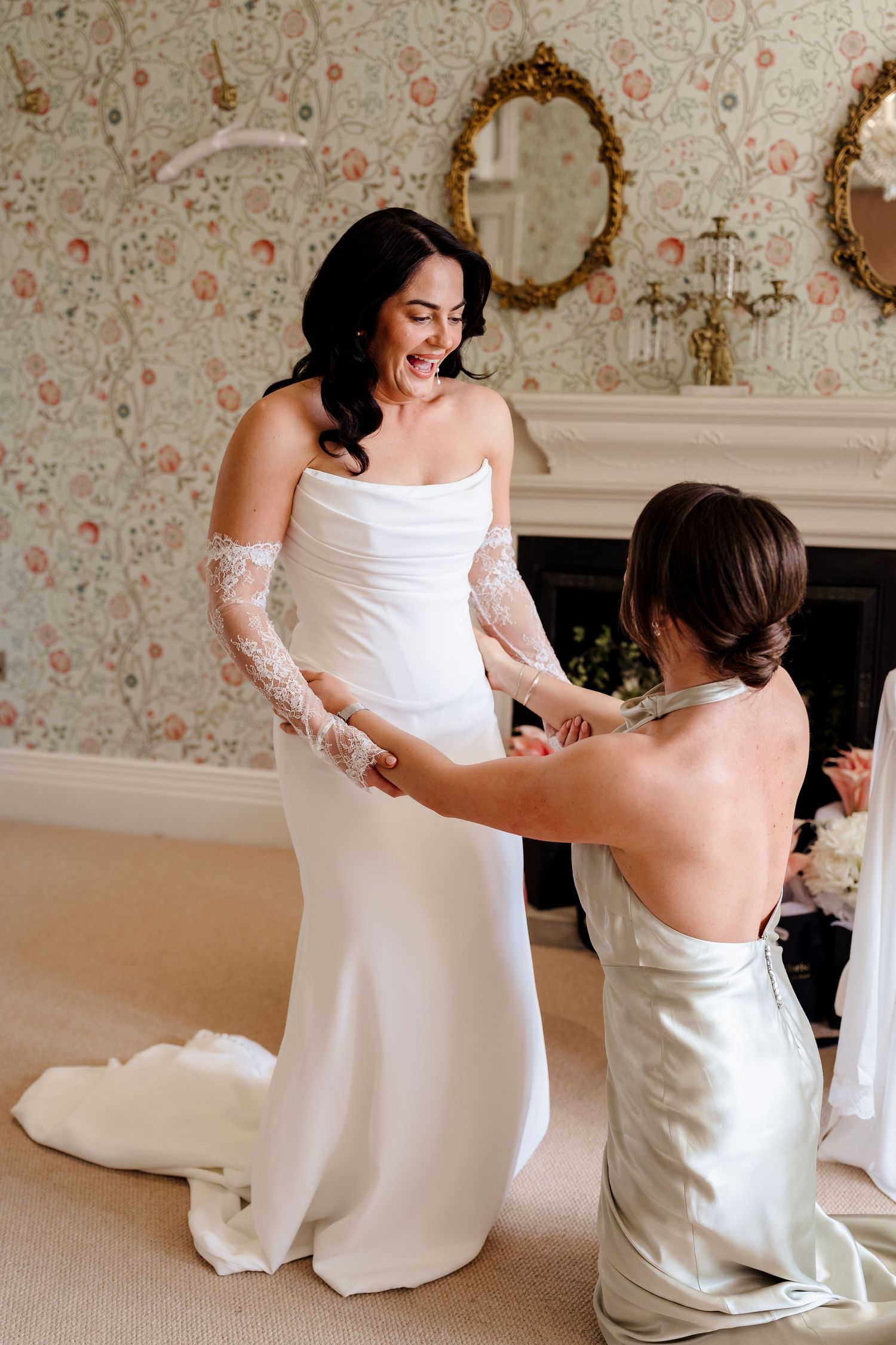 Bride smiling in elegant white gown with lace gloves as bridesmaid adjusts dress in floral wallpapered room.