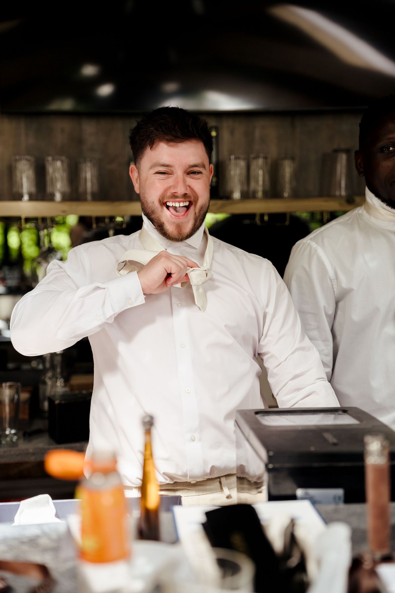 Laughing groom in white shirt adjusting his bow tie at a bar during wedding celebrations.