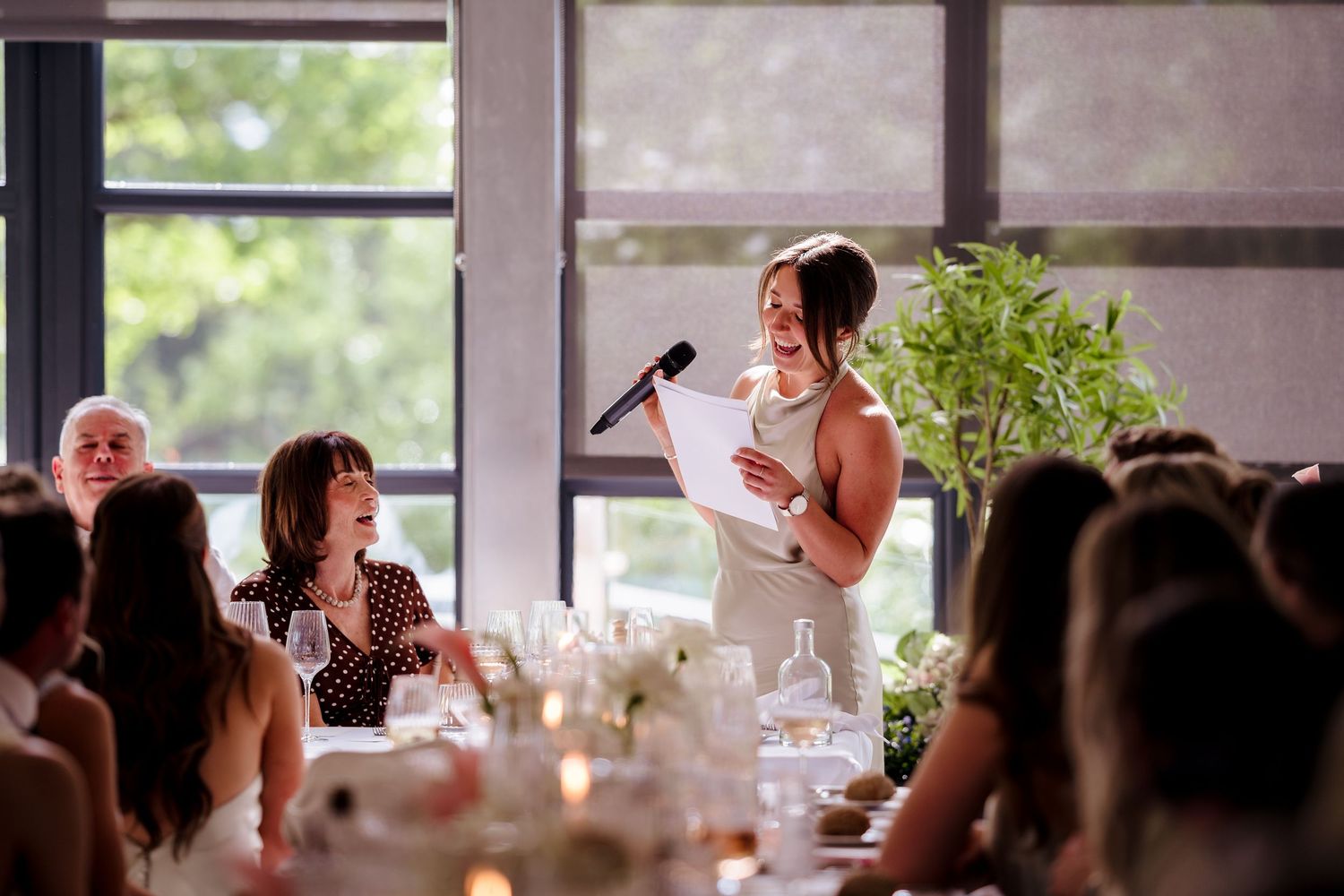 Woman giving a speech with microphone at an elegant wedding reception dinner, guests smiling and listening at decorated tables.