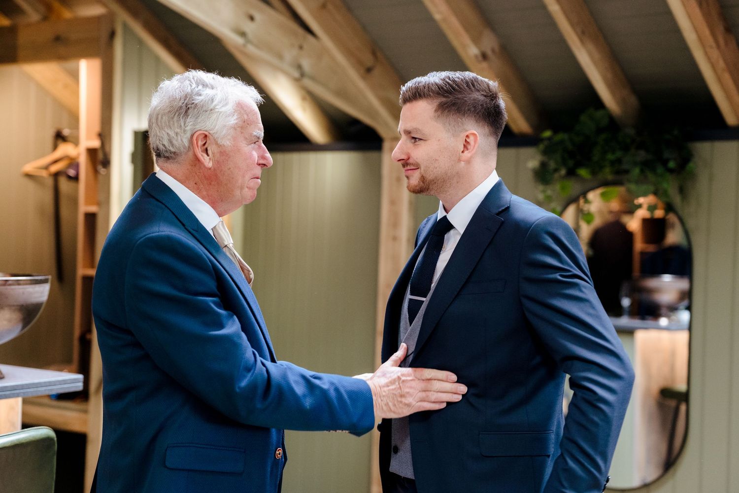 Two men in navy suits shaking hands and conversing in a rustic interior setting.
