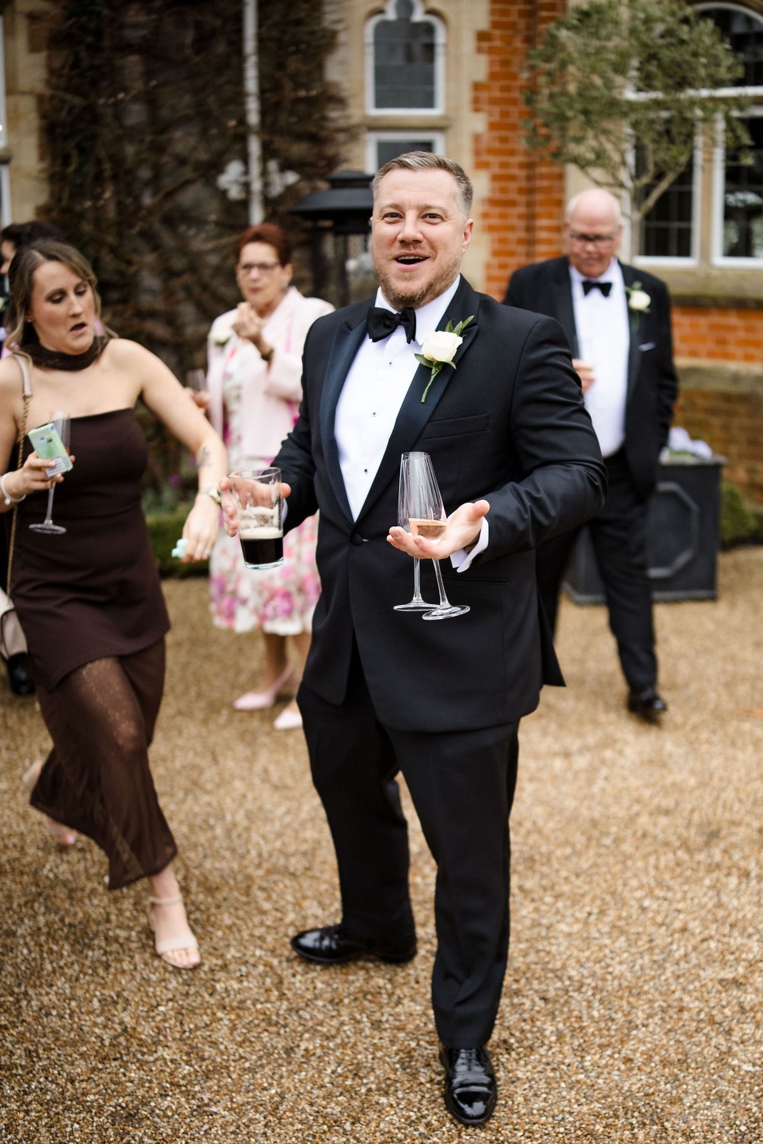 Smiling wedding guest in black tuxedo carries champagne glasses at an outdoor reception.