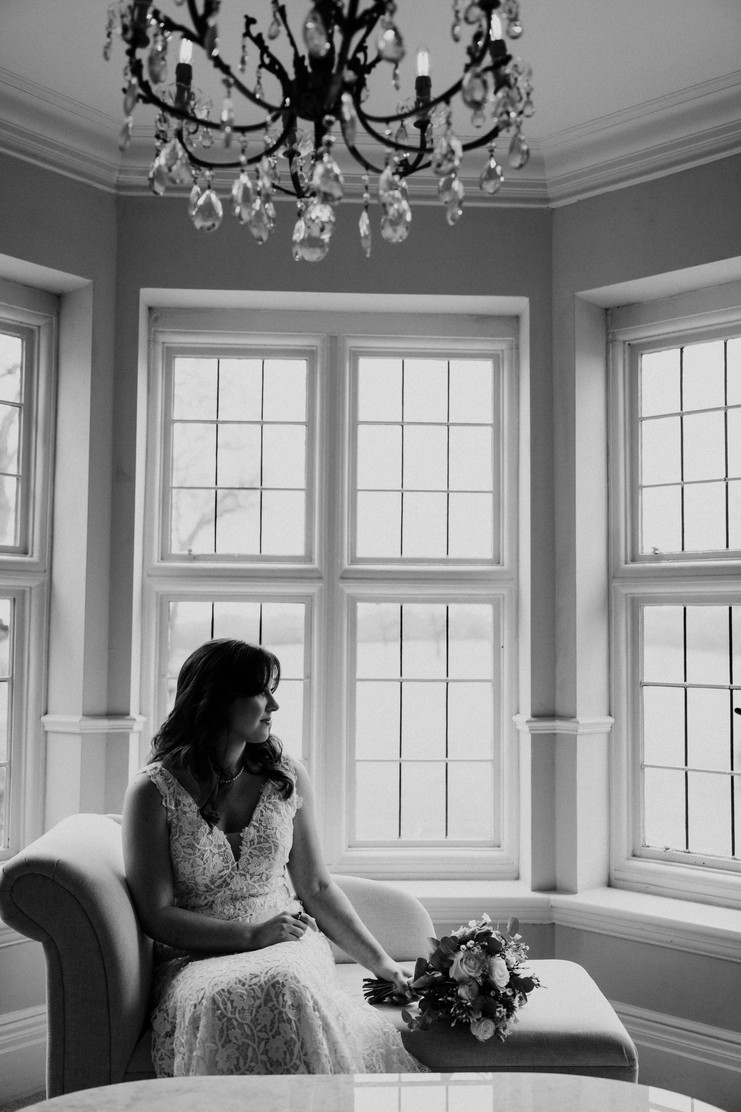 Bride in lace gown sits by bay window under chandelier in black and white portrait.
