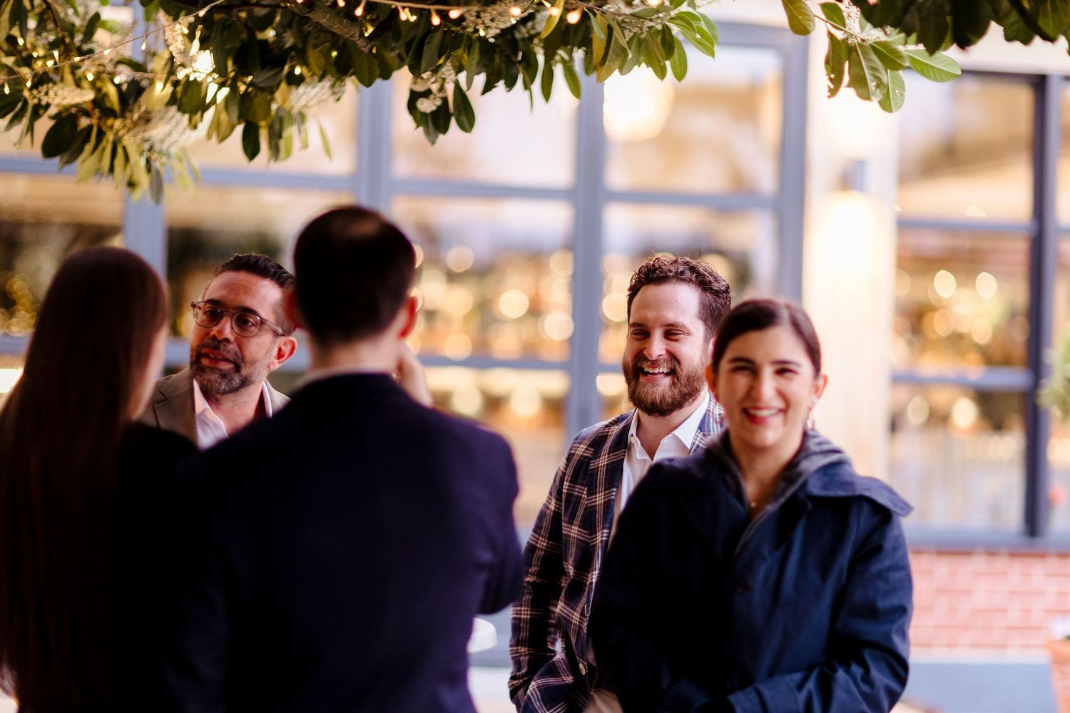 Smiling wedding guests mingle outdoors under leafy branches near illuminated venue windows.