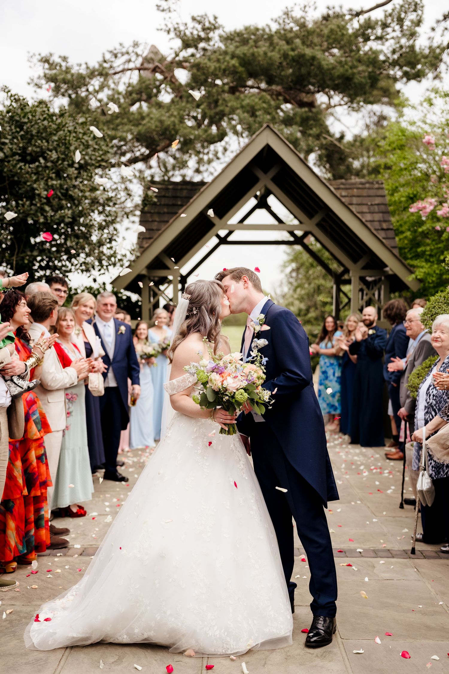Bride and groom share a kiss while guests throw confetti under a wooden arch at an outdoor wedding ceremony.