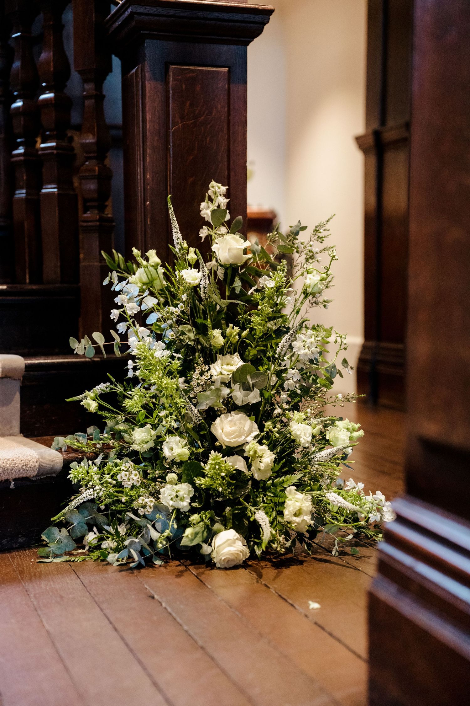 Elegant white floral arrangement with roses and greenery placed on wooden church floor beside dark wood staircase.