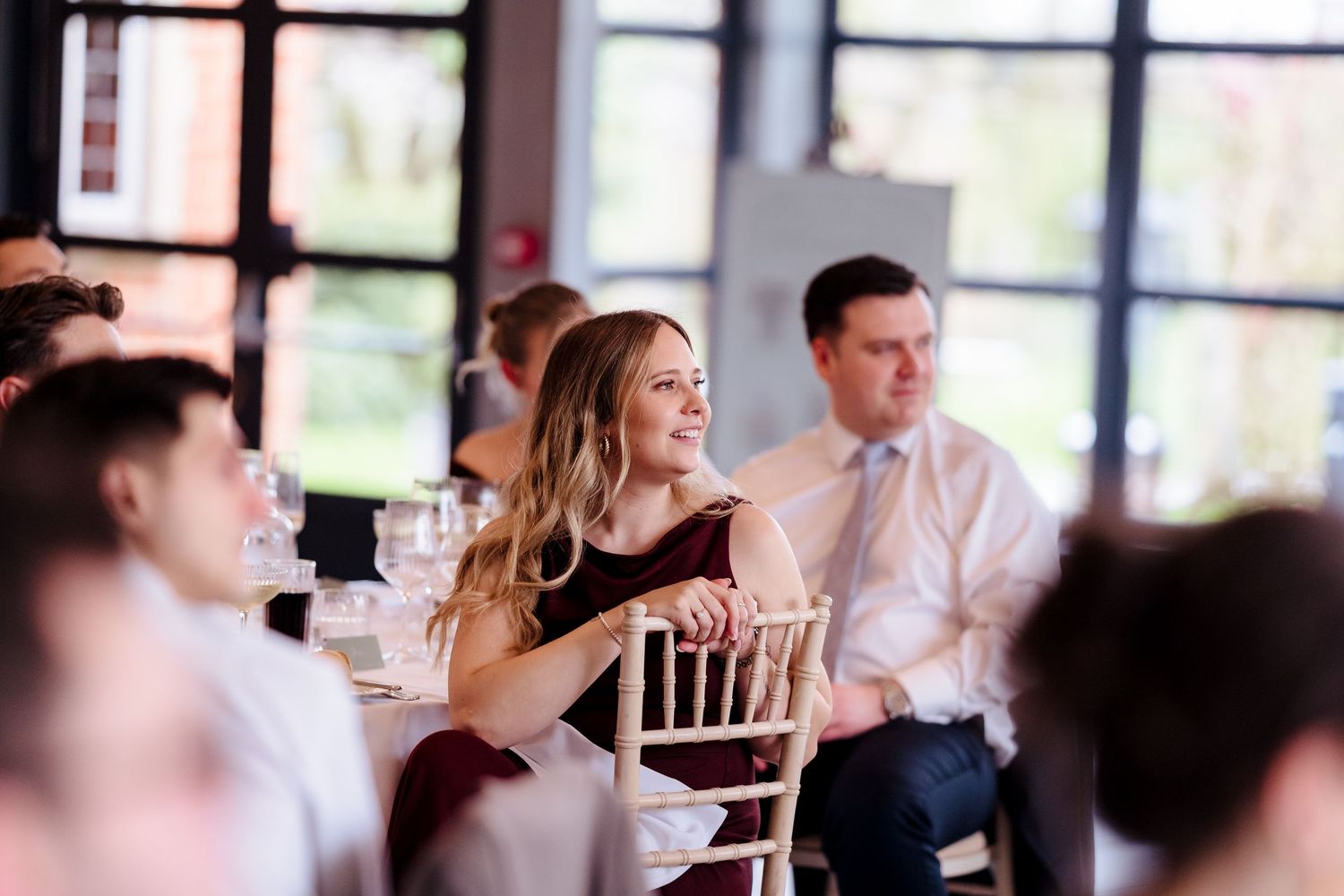 Smiling female wedding guest in burgundy dress seated at reception table listening to speeches.