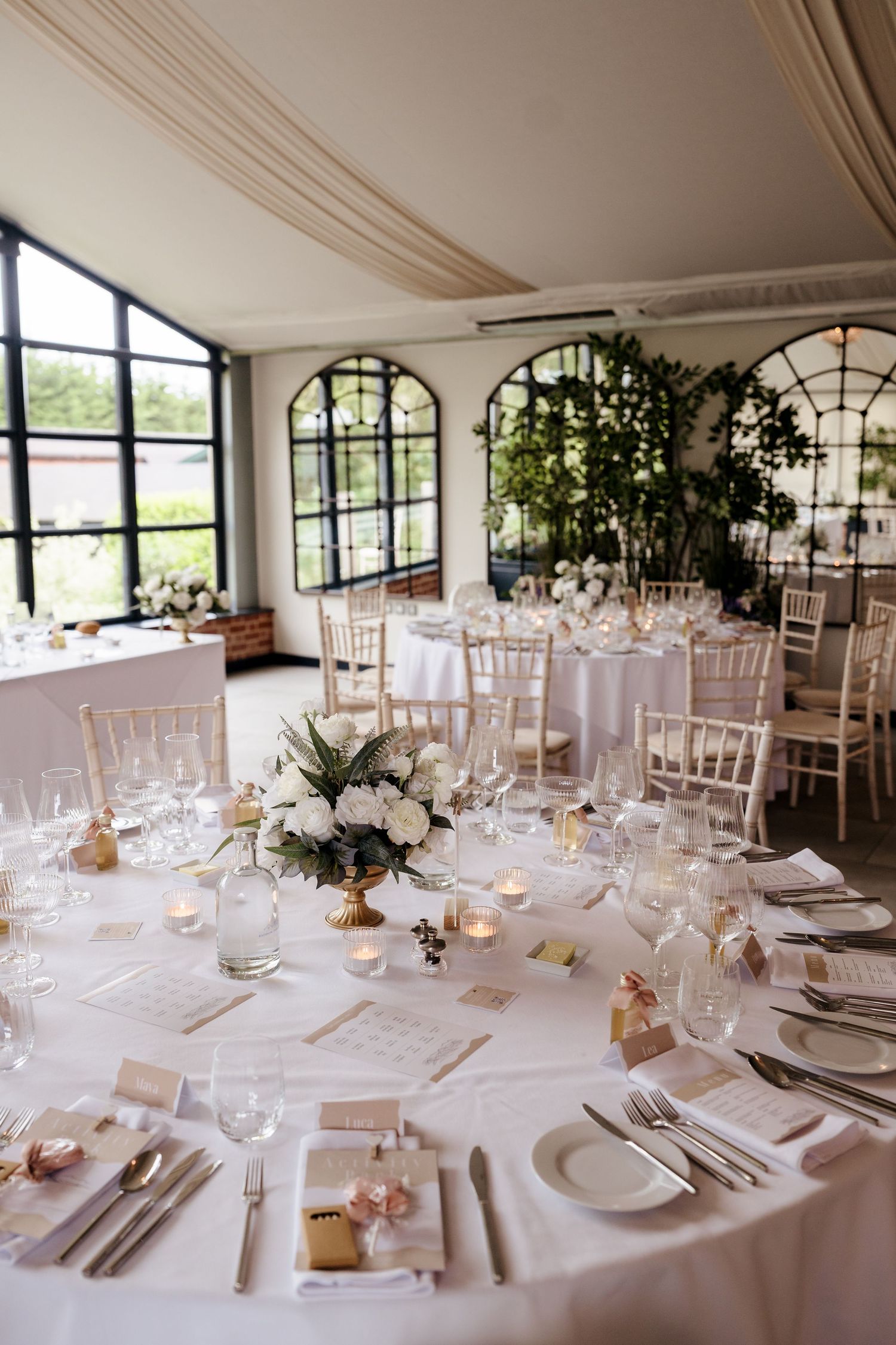 Elegant wedding reception hall with white round tables, floral centerpieces, gold chairs, and arched windows.