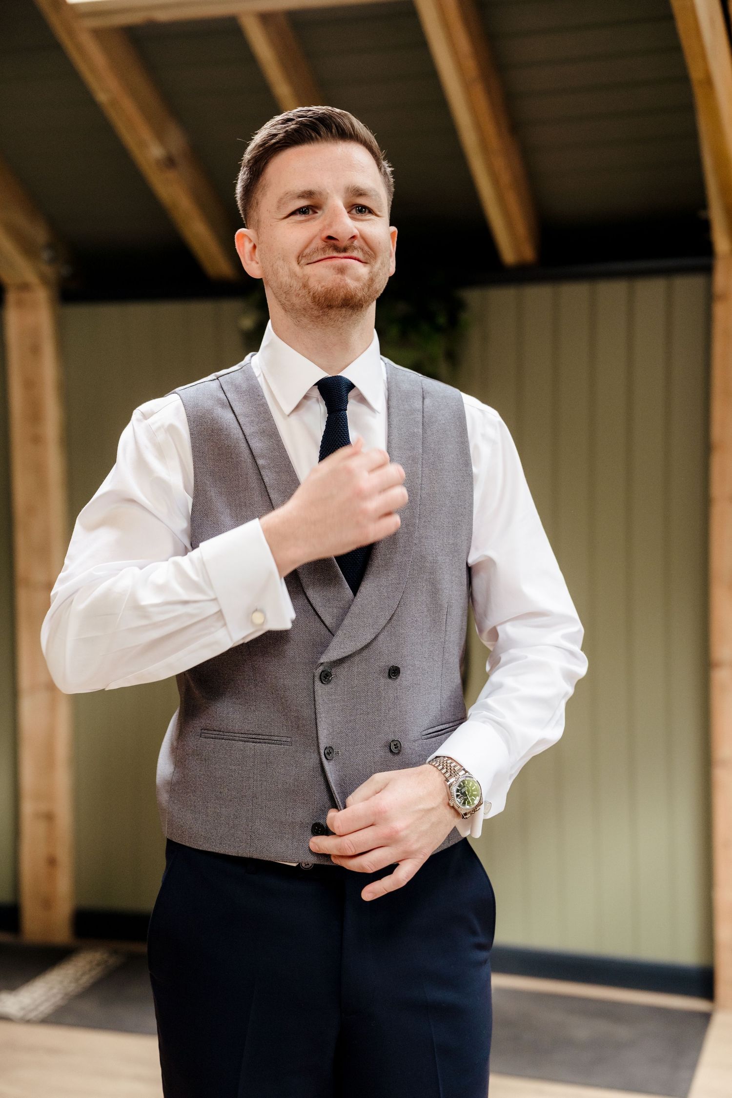 Smiling groom adjusting his tie wearing a grey waistcoat, white shirt, and dark trousers outdoors.