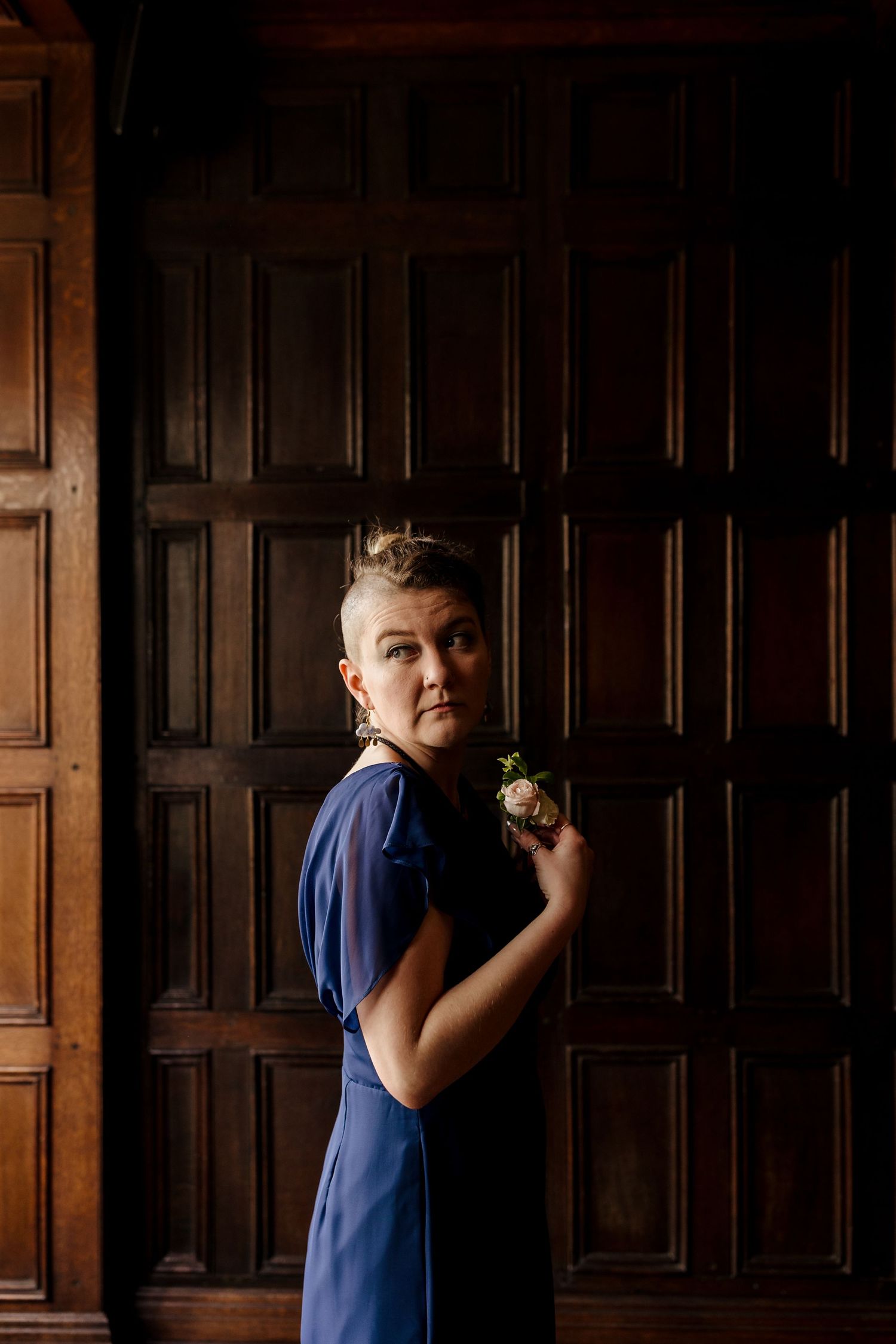 Woman in navy blue dress holding a boutonniere in front of ornate wooden paneled doors.
