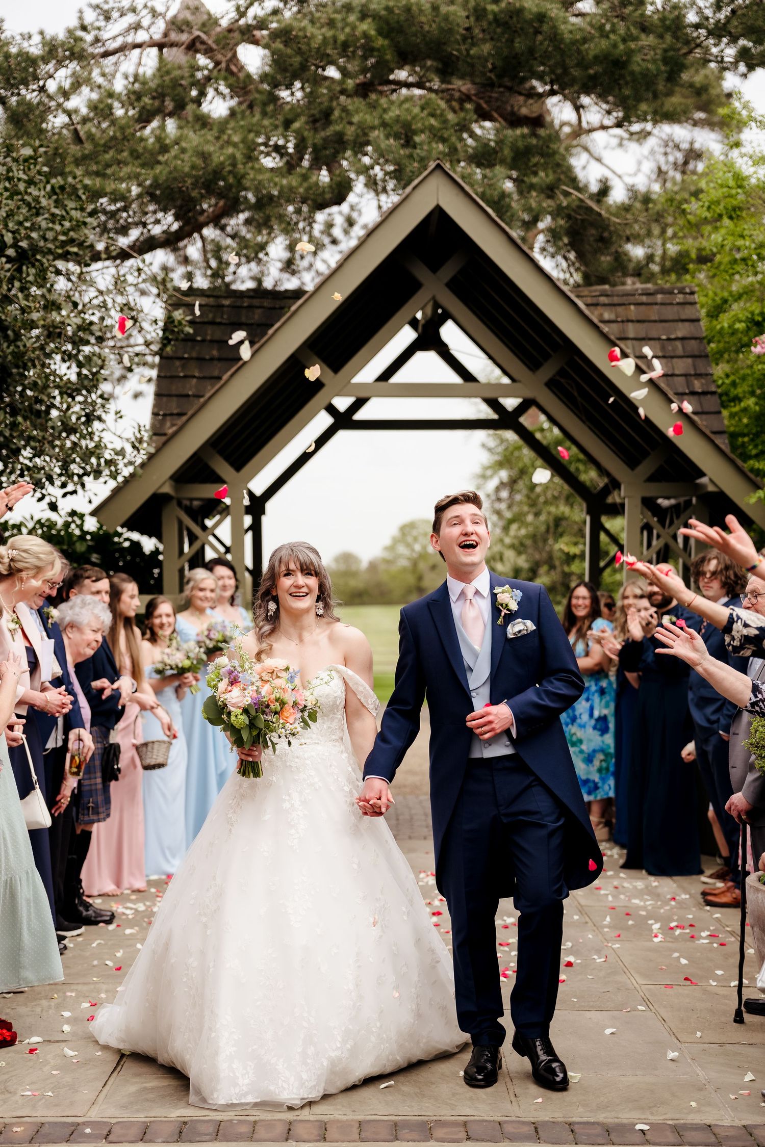 Joyful newlyweds walk through confetti shower under wooden arch, guests cheering in background.