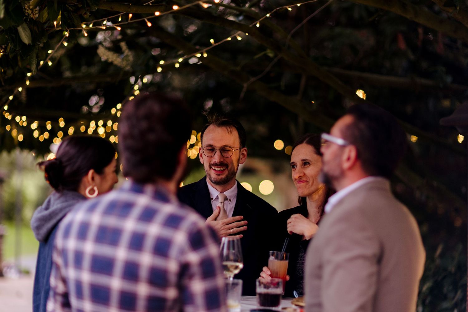 Wedding guests socializing under fairy lights in an outdoor evening reception setting.
